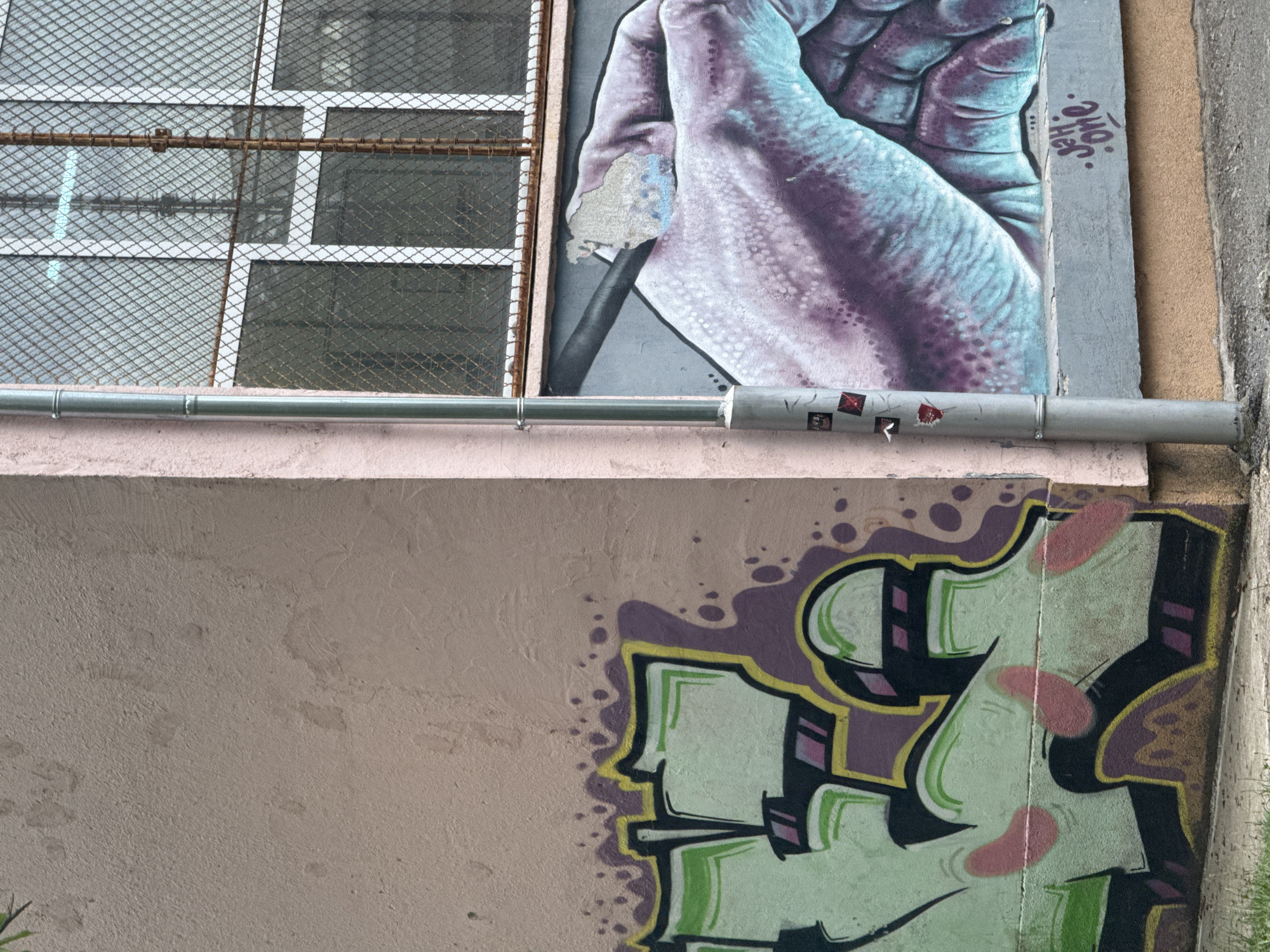 The image shows a close-up view of a building's exterior wall in Sofia, Bulgaria, featuring graffiti art. On the left side of the frame, a portion of a pink-colored wall displays a green and purple graffiti tag with yellow and black outlines. The graffiti appears to be abstract lettering. To the right of this, a downspout runs vertically along the wall, which transitions to a lighter pink color. Adjacent to the downspout is a section of a wall painted with a large mural depicting a human hand rendered in shades of purple and turquoise. The hand is drawn with detailed shading and texture, and appears to be holding a dark object, possibly a pen or pencil. The mural is partially damaged in the upper right quadrant, revealing a lighter colored surface underneath. Above the mural, a window with diamond-shaped metal grating is visible. The window has a rusty metal frame and appears to look into an interior space. The ground in front of the wall is visible, with some grass and concrete. Small stickers are affixed to the downspout. A signature, "Jah.one.", is spray-painted in purple graffiti below the hand mural.