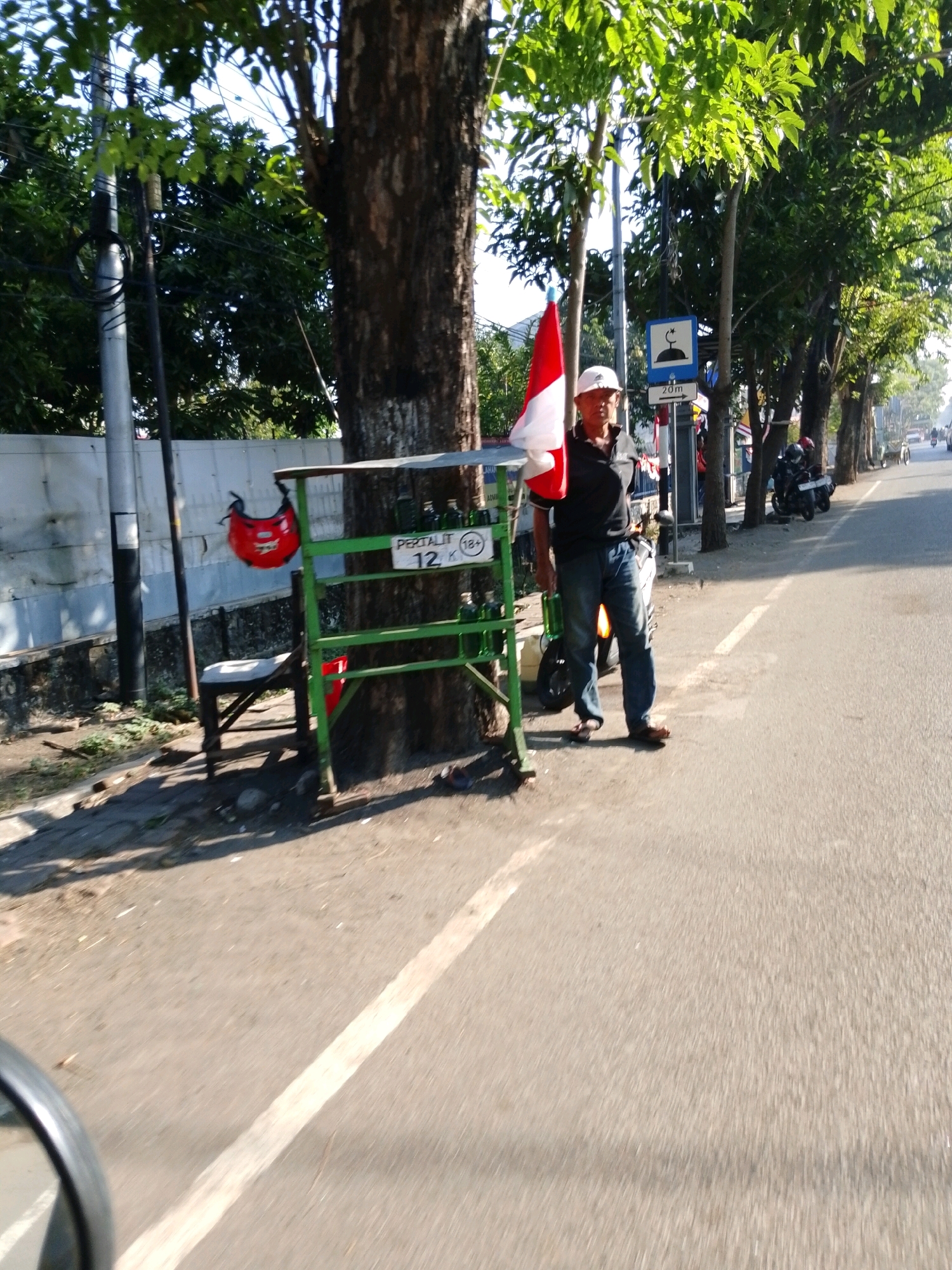 In the city of Pasuruan, Indonesia, a man stands on the side of a paved road, holding a red and white flag, likely the Indonesian flag. He is wearing a white cap, a dark polo shirt, and blue jeans. Next to him is a green metal stand with bottles on display and a sign that reads "PERTALIT 12 K". A red helmet hangs from a pole nearby, and a wooden chair sits behind the stand. Trees line the road, casting shadows on the ground, and a sign with a mosque symbol and "20m" is visible on a pole. In the background, motorcycles are parked along the roadside, and traffic can be seen further down the street. The scene appears to be a roadside stall selling fuel.