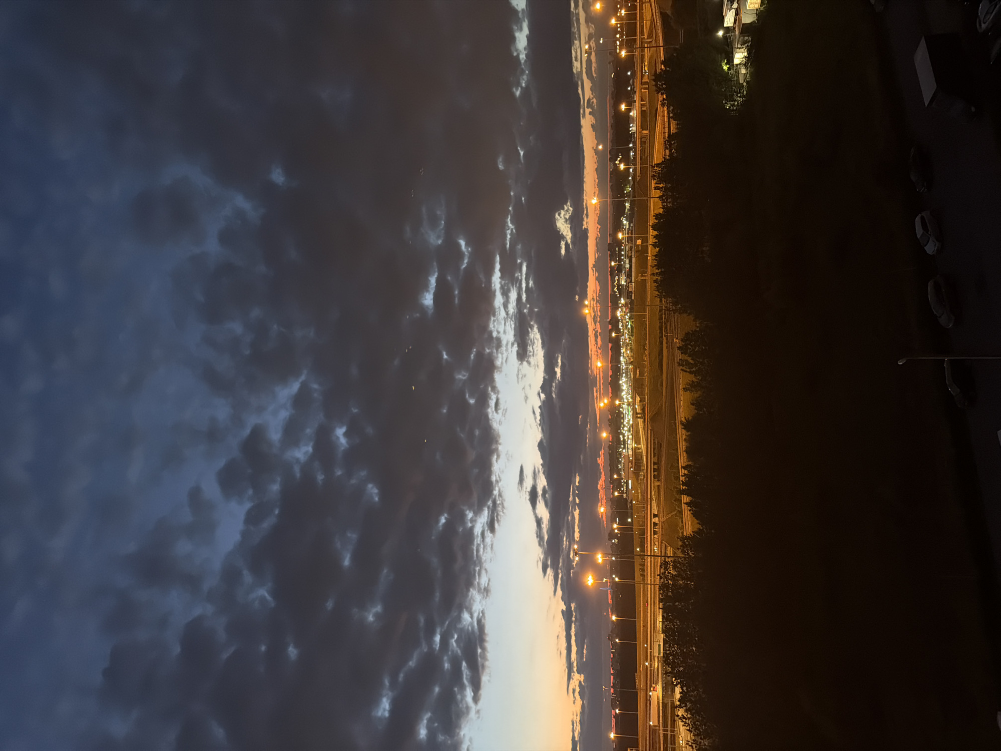 An exterior, vertical, medium shot captures an airport or industrial area at dusk under a cloudy sky. The sky is a gradient of dark blue at the top, transitioning to shades of gray and tinged with orange and yellow near the horizon, indicating the setting sun. Dark, textured clouds dominate the upper half of the frame.
Below the sky, a dense line of dark trees forms a silhouetted barrier across the middle ground. Beyond the trees, an expansive network of roads and infrastructure is illuminated by numerous streetlights, casting a warm glow. The road system features multiple lanes and overpasses, suggesting a busy transportation hub. In the distance, more lights indicate a built-up area.
The foreground consists of a dark, silhouetted area, possibly a building or elevated viewpoint. At the very bottom of the frame, a dimly lit area shows several parked cars and a dark rectangular structure, likely a truck or container. The scene is devoid of any visible people. The overall impression is one of urban development at the end of the day.
