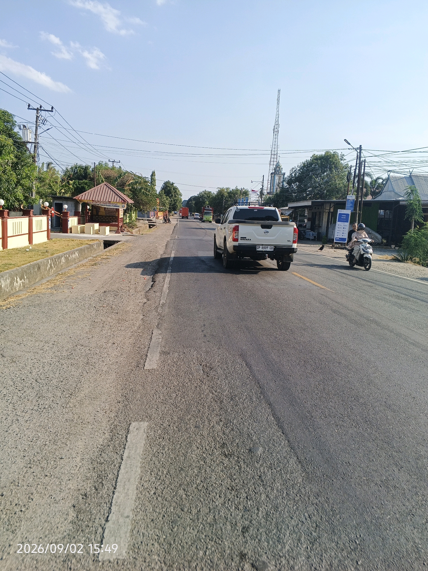 The image depicts a street scene in Pinrang, Indonesia, under a clear blue sky with scattered clouds. The road is paved with asphalt and marked with a yellow centerline and a white lane divider on the right side. On the left side of the road, there is a sidewalk area with a low concrete wall and a paved path leading to a small building with a reddish-brown roof. This building appears to be a public information booth or shelter, with signage visible. Adjacent to this, a beige fence with red accents encloses a grassy area.
In the foreground, a white pickup truck with the license plate "DP 8889 GB" is driving away from the camera. Further down the road, a white scooter carrying two people, one driving and one passenger wearing a headscarf and light-colored clothing, is also moving in the same direction. A black bird is visible on the roadside near the scooter.
On the right side of the road, there are several buildings with corrugated metal roofs, suggesting commercial or residential structures. Overhead, power lines and utility poles are prominent. In the background, a tall communication tower stands against the sky, along with trees and other vegetation. A red truck is visible in the distance, along with other vehicles. The overall impression is of a typical urban or semi-urban street in a tropical climate, with evidence of daily life and infrastructure. The timestamp "2026/09/02 15:49" is superimposed on the lower left corner of the image.