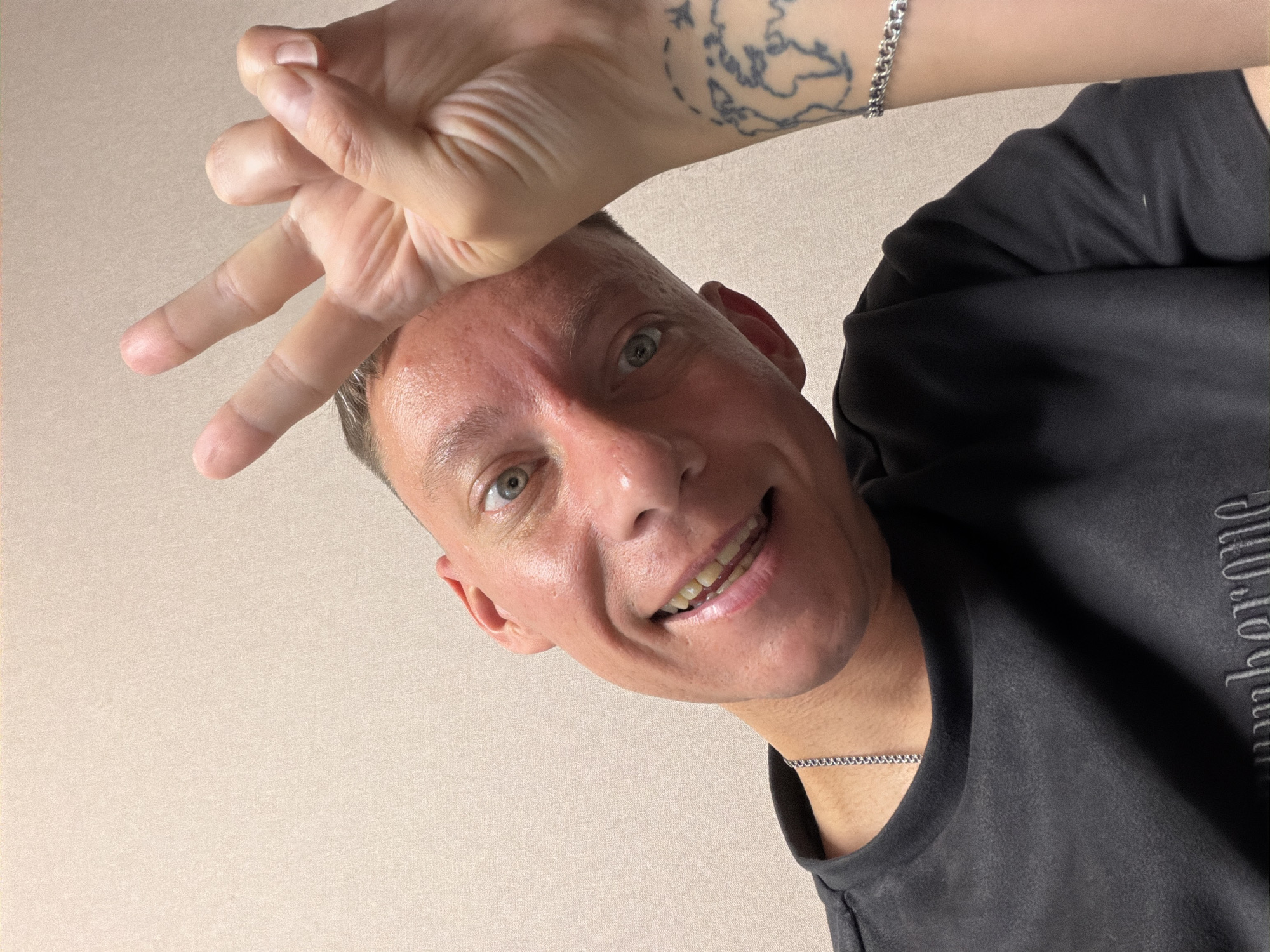 The image is a close-up, low-angle selfie of a fair-skinned person with short hair, looking directly at the camera and smiling. The person's right hand is raised, forming a peace sign in front of their face. They are wearing a dark gray t-shirt with a chain necklace visible. A tattoo of a world map is visible on their left forearm, along with a silver bracelet. The background is a plain, light-colored wall. There is no indication of the location being in Moscow, Russia.