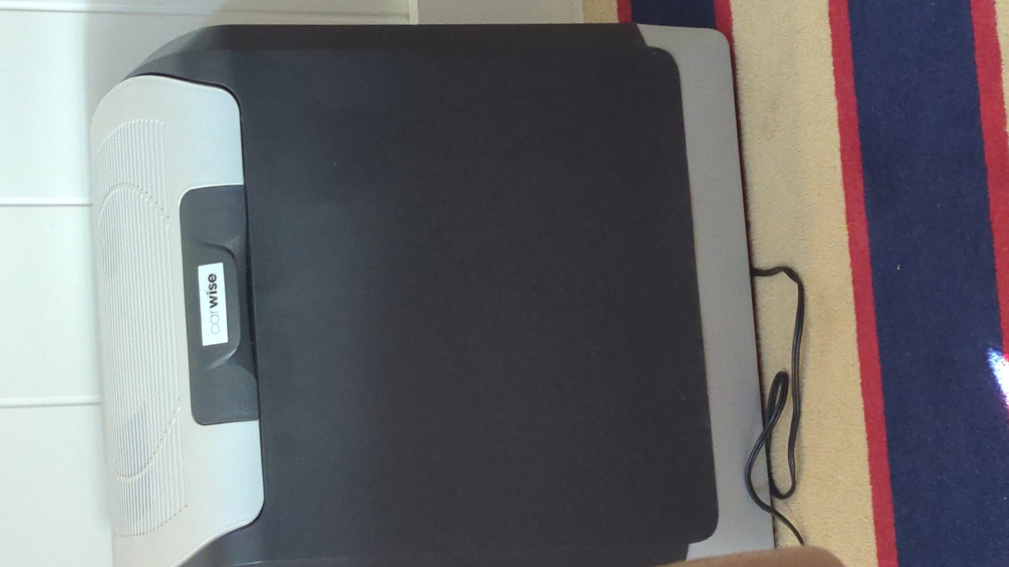 A black and gray portable cooler with a "carwise" logo is positioned on a carpeted floor. The cooler has a gray lid with a textured pattern and a dark gray front panel. A black power cord is plugged into the back of the cooler and lies on the carpet. The carpet has red, blue, and cream stripes. The background appears to be a white, paneled wall. The scene is indoors, and no specific location in Larkollen, Norway, is discernible from the image.