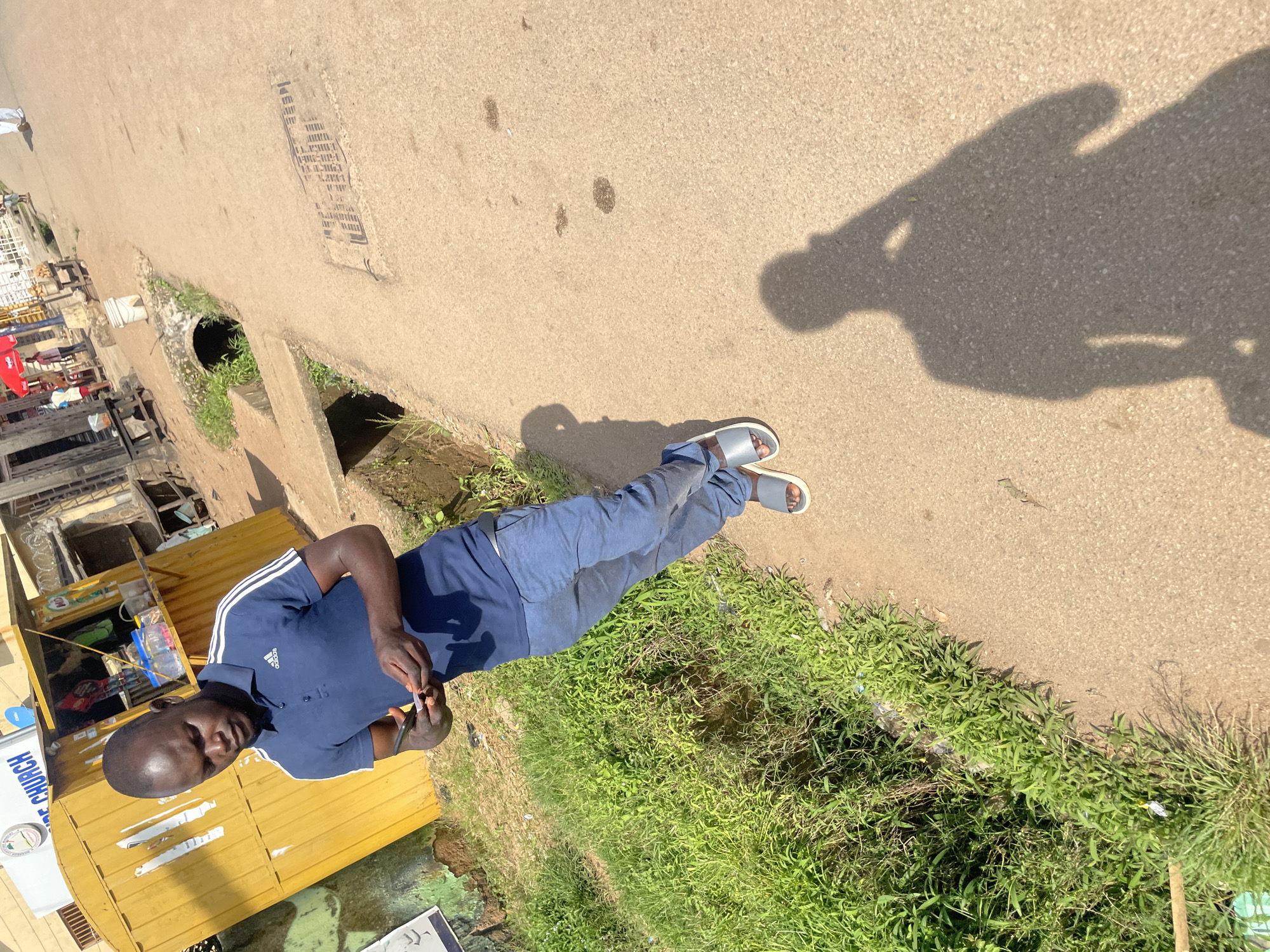 A man stands on a paved road in Jos, Nigeria, looking down at his phone. He is wearing a dark blue Adidas polo shirt and blue pants. He has on grey sandals. To his left is a yellow kiosk with signage that reads "CHURCH." The kiosk appears to be a small business. There is overgrown green vegetation to the left of the road and a drainage ditch. The road has a manhole cover and some dark patches, possibly from water. The man's shadow is cast on the road to his right. In the background, there are buildings and other people walking, some associated with market stalls.