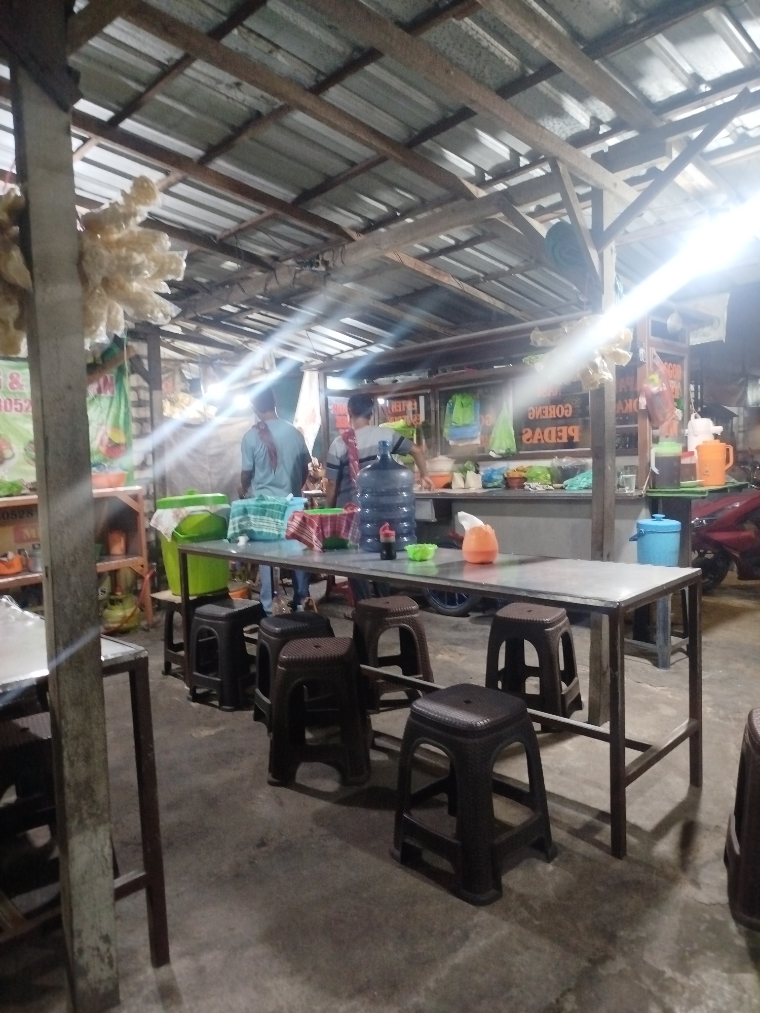 The image depicts an outdoor eatery located in Tonjung, Indonesia. The scene is characterized by a rustic, open-air structure with a corrugated metal roof supported by wooden beams. Underneath the roof, a long, metal-topped table is set with several plastic stools arranged around it. On the table, there are various items including a large water dispenser, a blue bucket, a tissue dispenser, and some food containers. Behind the table, a counter area is visible with more food items, containers, and what appears to be cooking equipment. Two individuals, presumably staff members, are working behind the counter, one of whom is seen from the back. In the background, a red motorcycle is parked, suggesting it belongs to a customer or staff member. The overall impression is that of a casual, local dining establishment.