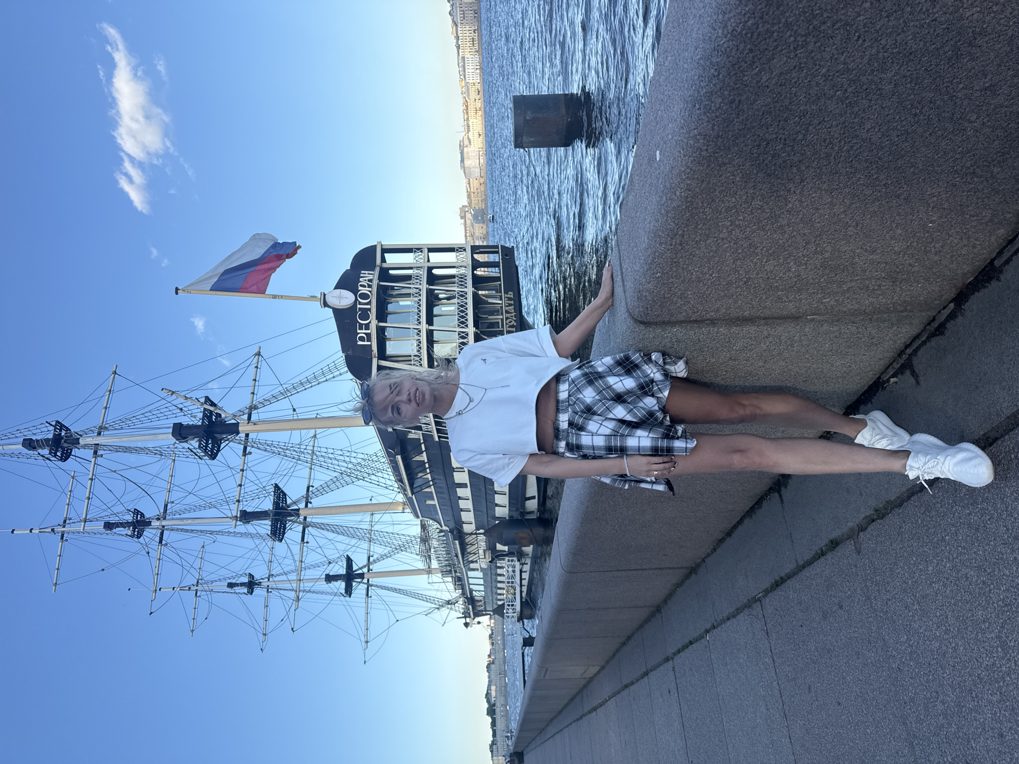A young woman with blonde hair is standing on a stone embankment next to a large, dark, multi-masted ship. The ship has a Russian flag flying from its mast and the word "РЕСТОРАН" (Restaurant) visible on its side. She is wearing a white crop top, a black and white plaid skirt, and white sneakers. The embankment is paved with concrete and has a granite wall. The background shows a body of water, likely a river or canal, and buildings on the opposite shore, characteristic of Saint Petersburg, Russia. The sky is blue with a few wispy clouds.