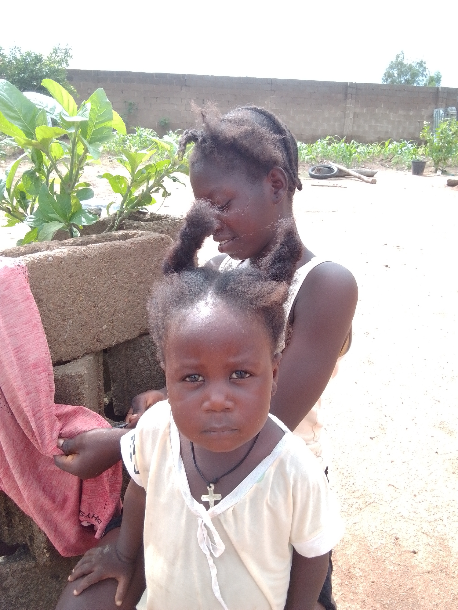 Two young girls, one older than the other, are posing for a photograph outdoors. The younger girl is in the foreground, facing the camera directly, with a neutral expression. She is wearing a pale yellow t-shirt with a white trim and a cross pendant on a black string. The older girl is behind her, her head tilted slightly to the side. She has dark braided hair that is pulled up into two pigtails. A pink cloth is draped over a stack of concrete blocks to the left of the frame. The background suggests a rural or semi-urban setting with a brick wall, some greenery, and a sandy ground. The sky is bright and overcast, indicating daylight. The overall impression is of a candid moment captured in a humble outdoor environment, likely in the village of Howayi, Nigeria.