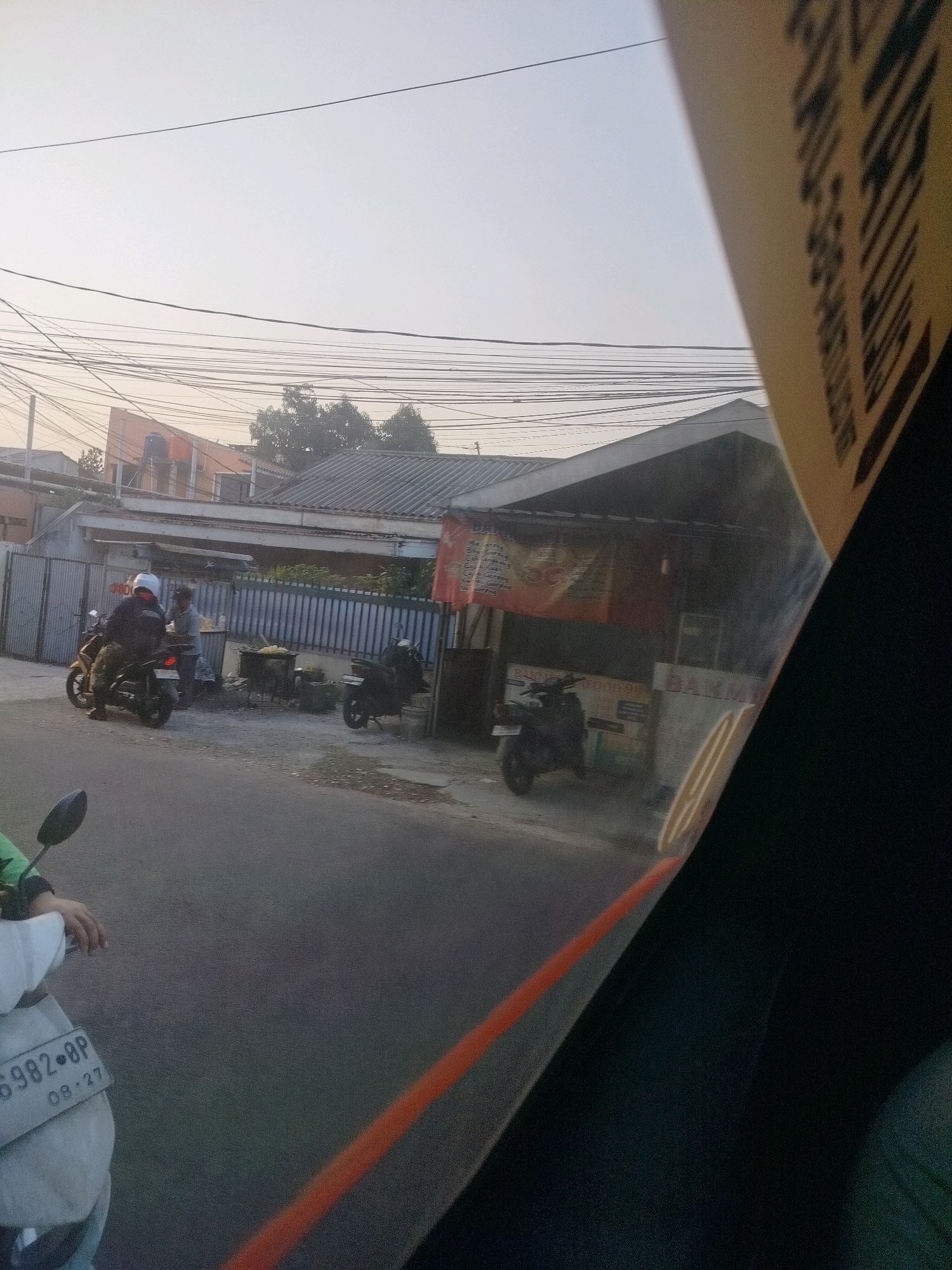 The image depicts a street scene in East Jakarta, Indonesia, possibly during the late afternoon due to the hazy sky. In the foreground, a portion of a vehicle, likely a car or motorcycle, is visible, with a yellow label bearing text in Indonesian and a distinct orange line on its surface. The street is paved and shows some debris. On the right side of the road, a food stall is set up with several individuals present. One person on a motorcycle wearing a white helmet is arriving at the stall, while another person appears to be working at the stall, possibly serving customers. A second motorcycle is parked near the stall. The background features a row of buildings with corrugated metal roofs, typical of residential or commercial structures in the area. Numerous electrical wires crisscross overhead, a common sight in urban Indonesian landscapes. Some trees are visible behind the buildings. The overall atmosphere suggests a bustling everyday street life.