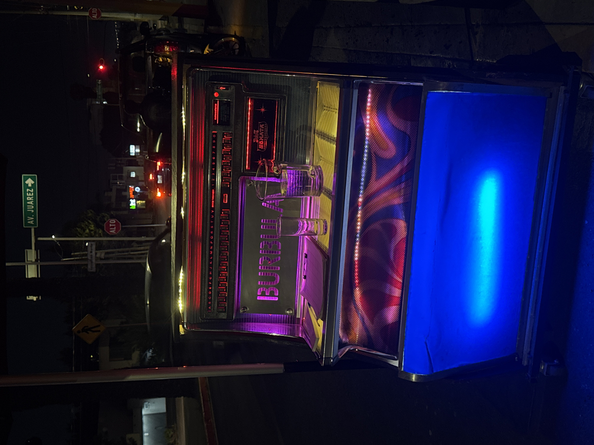 The image, captured at night in Puebla, Mexico, features a brightly lit, colorful jukebox as its central subject. The jukebox displays prominent red, purple, and blue lighting, suggesting an active use in a nighttime social setting. Its main panel includes rows of buttons for song selection, labeled with numbers 1 through 9 and 0, and letters A through Z. Visible text on the jukebox includes "Golesalana," "BURRILI," "SONATA," and "UV DISCO TOCANDO." Two clear drinking glasses are placed on top of the machine.
The background reveals a dark street scene with various urban elements. A green street sign clearly reads "AV. JUAREZ" with a directional arrow. Two red octagonal "ALTO" (STOP) signs are visible on street poles, one in the mid-left and another further back. A traffic light showing a red signal, along with other distant lights from buildings and vehicles, confirms the nighttime setting. A yellow diamond-shaped sign indicating a pedestrian crossing is also present. The scene depicts an active, illuminated outdoor environment, possibly a street-side establishment, where the jukebox serves as a source of entertainment.