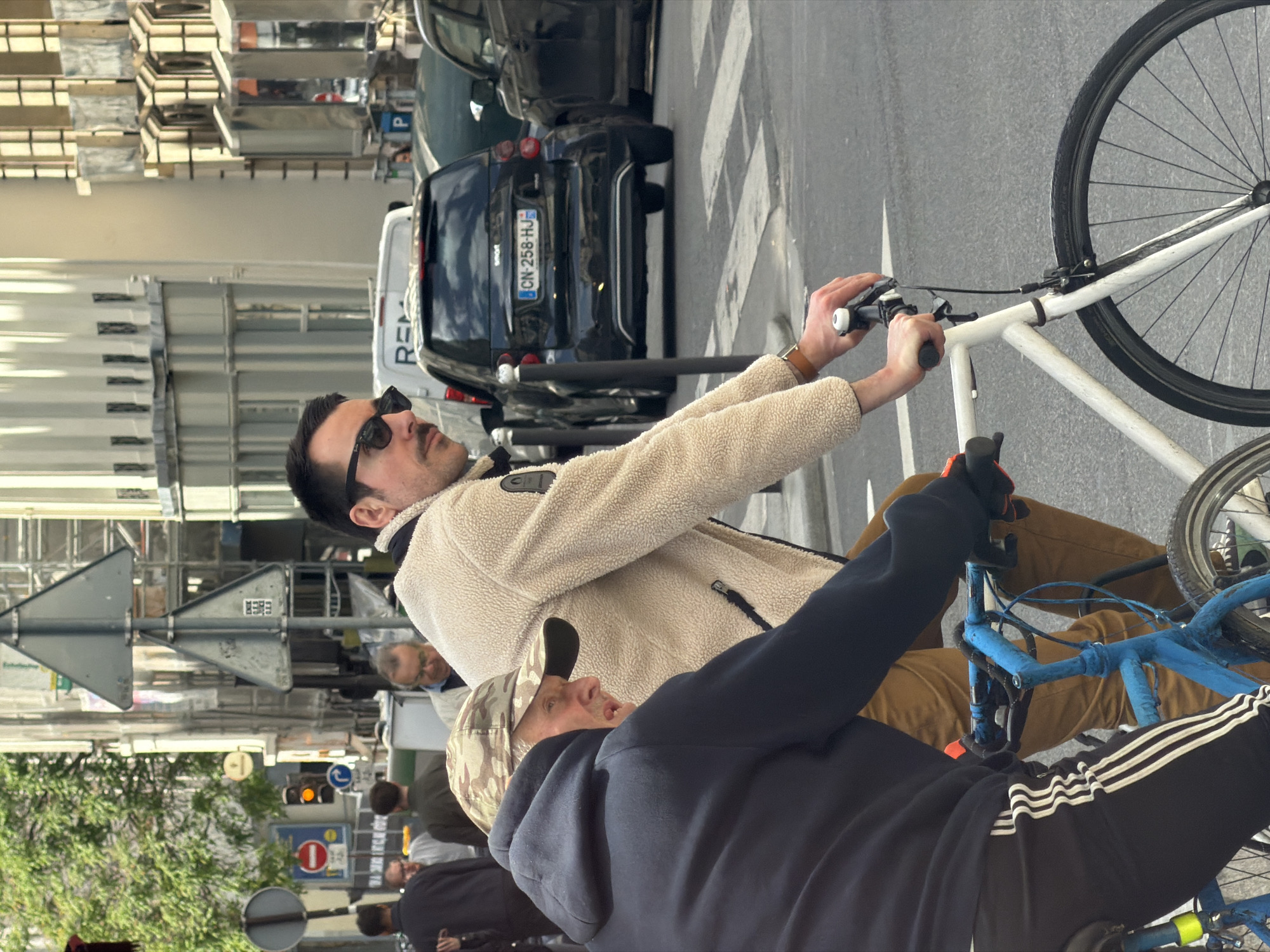 This image captures a daytime street scene in Paris, France, featuring two men on bicycles with urban surroundings. The perspective is slightly elevated, looking across the street.
In the foreground, a man wearing a camouflage baseball cap and a dark blue hoodie with white stripes on the sleeves is partially visible, positioned on a blue bicycle. He appears to be looking towards the second cyclist.
Behind him, another man with a mustache and dark sunglasses is prominent, wearing a cream-colored, textured jacket. He is seated on a white bicycle, holding the handlebars with both hands, suggesting he is actively riding or has just paused. His gaze is directed forward and slightly to his right.
The background reveals a typical city street environment. A black Smart car is parked along the curb, displaying the French license plate "CN-258-HJ". A white van with "REM" visible on its side is also parked further down. The street features a zebra crossing. Various street signs are present, including a triangular sign, a red circular "no entry" sign, and a blue circular mandatory direction sign, alongside traffic lights. Buildings line the street to the left, and green foliage from trees is visible in the lower left of the frame. The scene portrays a bustling urban setting.
