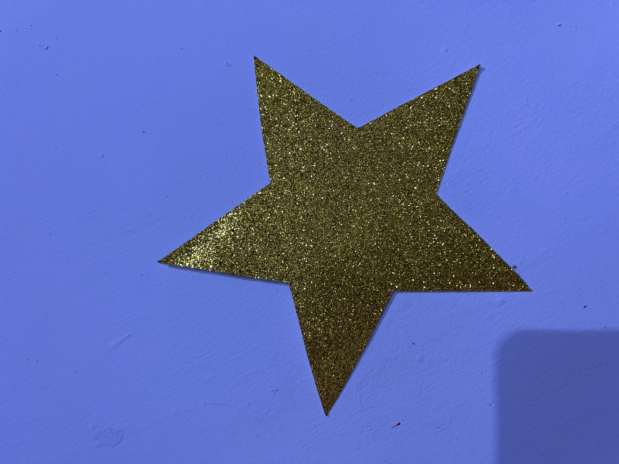 A golden glitter star decoration is affixed to a textured, light blue wall. The star is centrally located and oriented with one point facing upwards. The lighting casts a soft shadow to the lower left of the star. The wall appears to be an interior surface, possibly in a residential or decorative setting. There is no information provided about the location in Santa Ana, Venezuela.