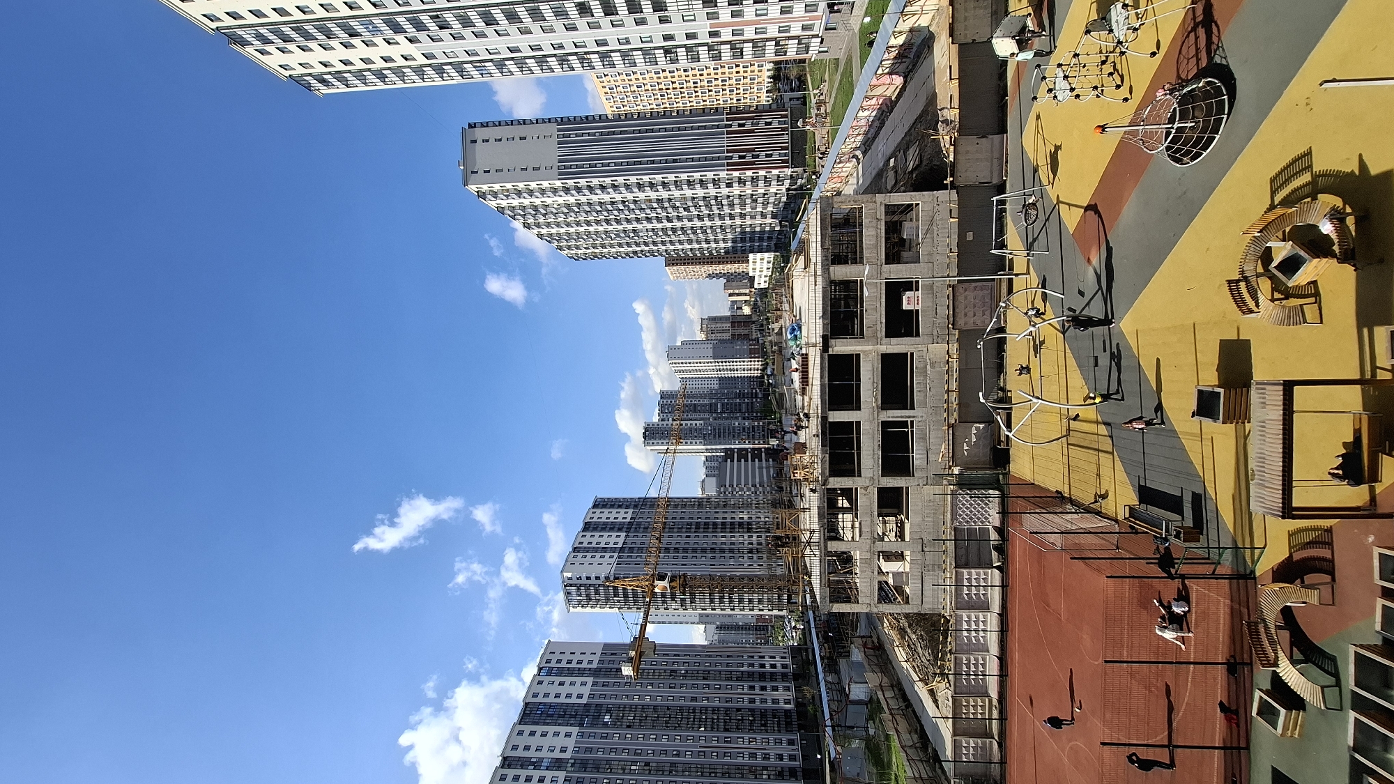 The image shows a construction site and surrounding buildings in what appears to be a dense urban environment. High-rise residential buildings of varying heights are visible in the background. In the foreground, a building is under construction, with its concrete frame exposed. A construction crane stands nearby. Adjacent to the construction site is a sports ground and children's playground. Several individuals are visible on the sports ground. The image appears to be taken from an elevated position. The city of Saint Petersburg, Russia, is suggested as the location.