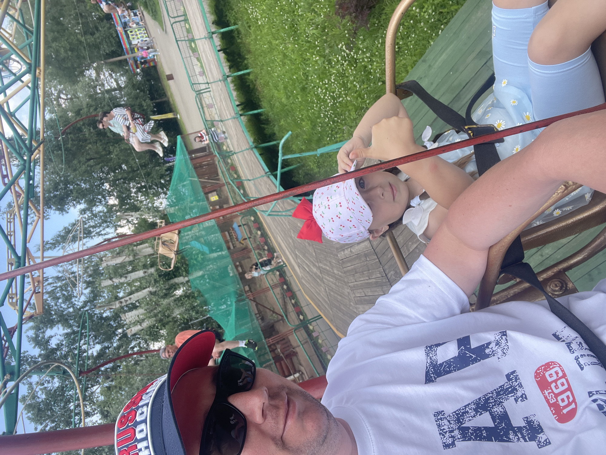 A father and daughter are on a ski lift ride in the city of Chaikovsky, Russia. The father is in the foreground, wearing a white t-shirt with a blue graphic and a red, white, and blue baseball cap. He is wearing sunglasses and looking towards the camera. The daughter is next to him, wearing a white cap with a red bow and a light blue dress with white daisy patterns. She is giving a thumbs-up to the camera. In the background, other people are visible on ski lifts, and there are trees, grass, and amusement park structures. The ski lift appears to be in an outdoor amusement park or recreational area.
