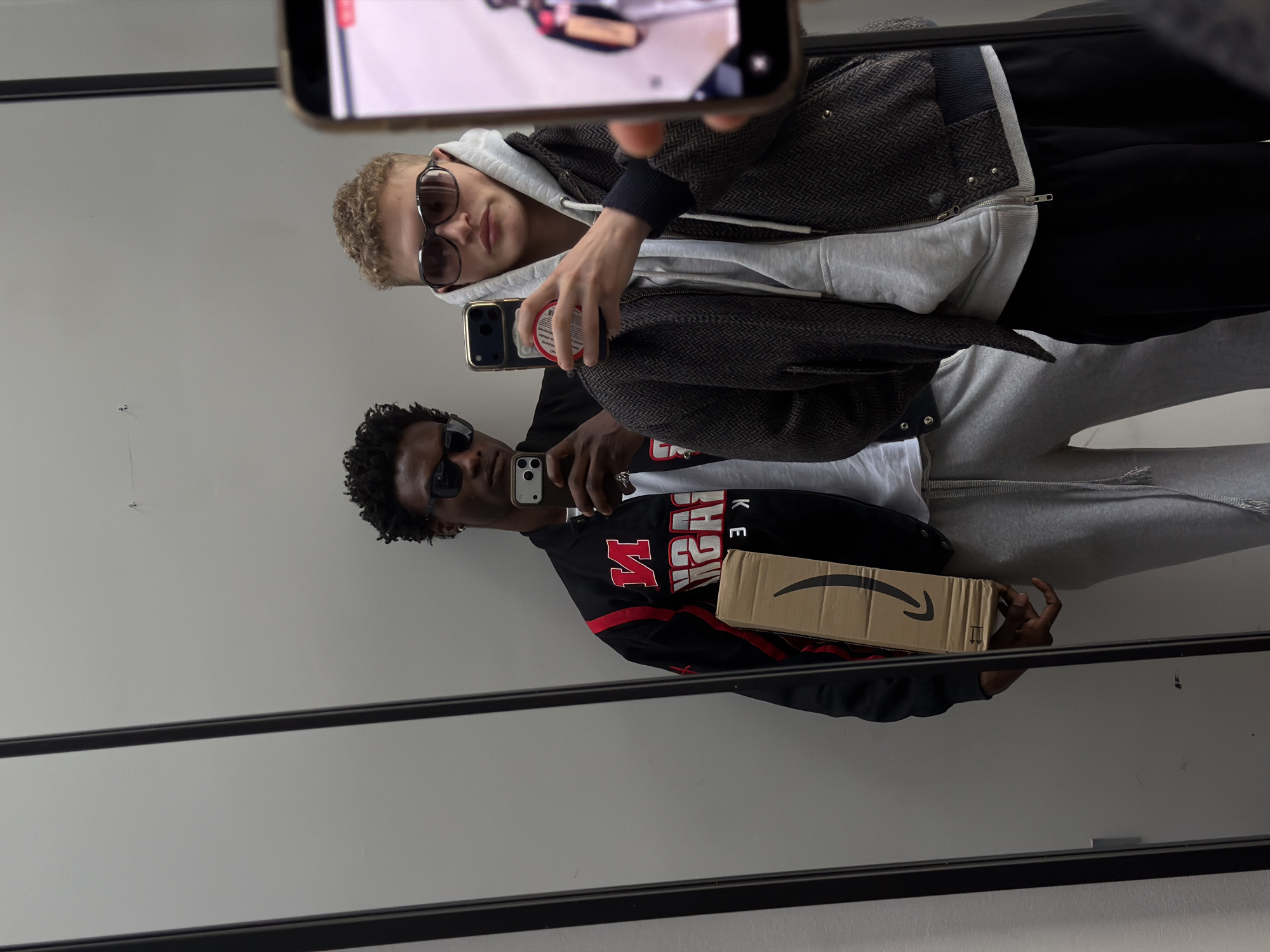 Two men are taking a selfie in front of a mirror. The man at the top has blond hair and is wearing sunglasses, a grey hoodie, and a brown jacket. He is holding a phone and has what appears to be another phone in his hand. The man at the bottom has dark hair and is wearing sunglasses and a jacket with the word "Nebraska" on it. He is holding a phone and an Amazon box. The background is a white wall with black trim. The location is not possible to determine from this image.