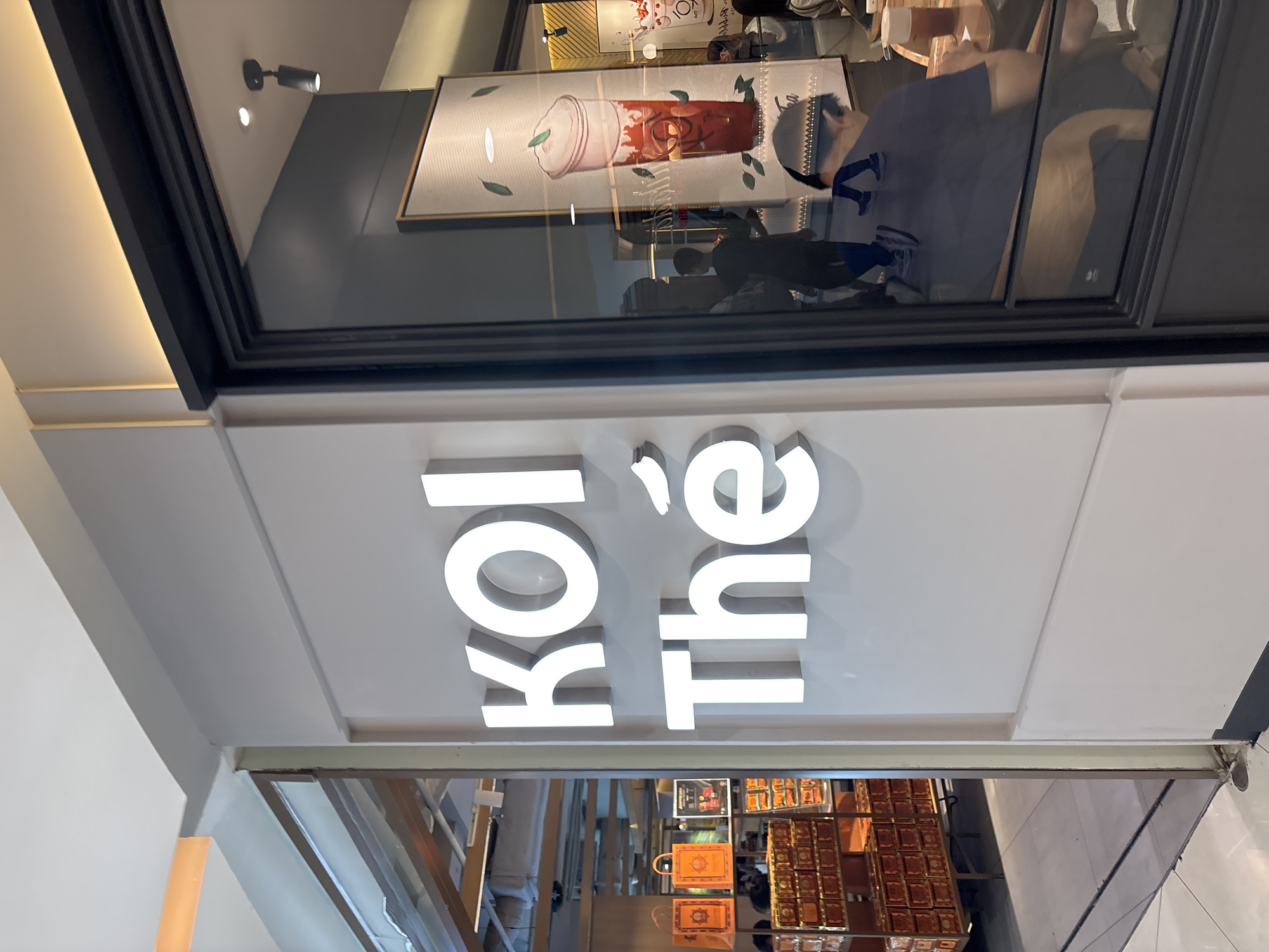 The image shows the exterior of a KOI Thé bubble tea shop. The shop's name is displayed in large, illuminated white letters on a grey wall. To the left of the shop's entrance, there are shelves filled with packaged products, possibly tea or gift boxes, and orange shopping bags. Inside the shop, visible through a large glass window, several customers are seated at tables. One person is seen from the back, wearing a dark blue t-shirt and sitting at a table with a drink. Other patrons are also visible in the reflection and inside the shop. The shop appears to be located in a mall or commercial area. This KOI Thé outlet is located at Neo Soho, located in the city of West Jakarta, Indonesia.