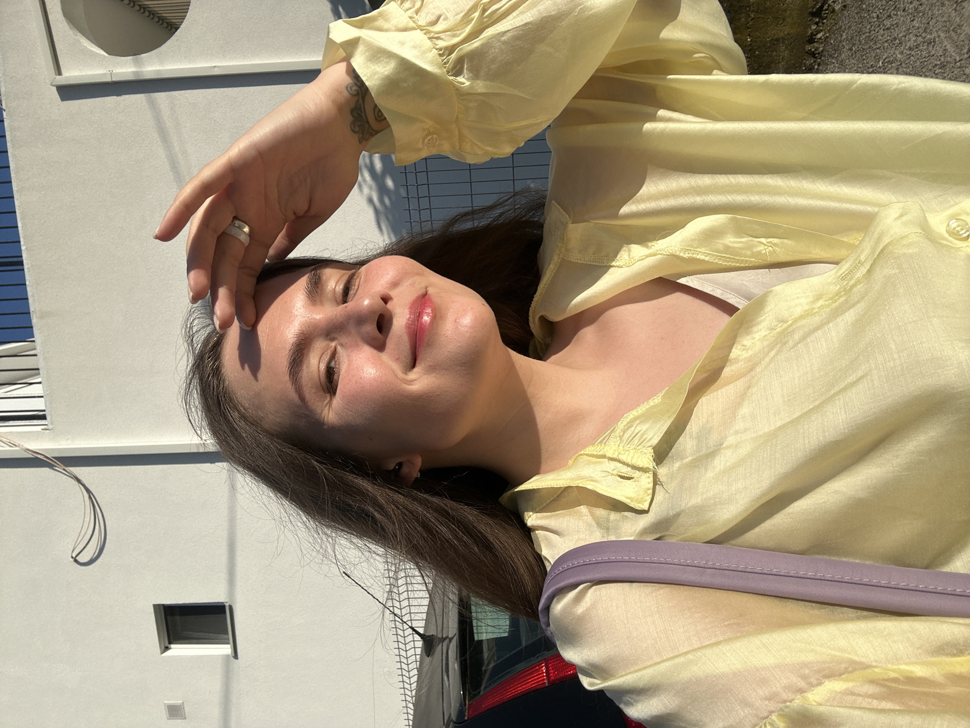A young Caucasian woman with long brown hair is taking a selfie outdoors. She is wearing a pale yellow, translucent blouse with puffy sleeves and a light-colored camisole underneath. A purple strap of a bag is visible on her left shoulder. Her right hand is raised, touching her forehead, and she is wearing a ring on her index finger and a tattoo on her wrist. She is smiling, and the sun is shining on her face, creating highlights.
The background consists of a white building with a small window and some wiring on the wall. A black car is partially visible on the left, and a metal fence is behind the woman. Shadows of the fence and possibly trees are cast on the ground and the building. The scene appears to be in a residential or commercial area in the city of Šušanj, Montenegro, suggested by the architecture and fencing.
