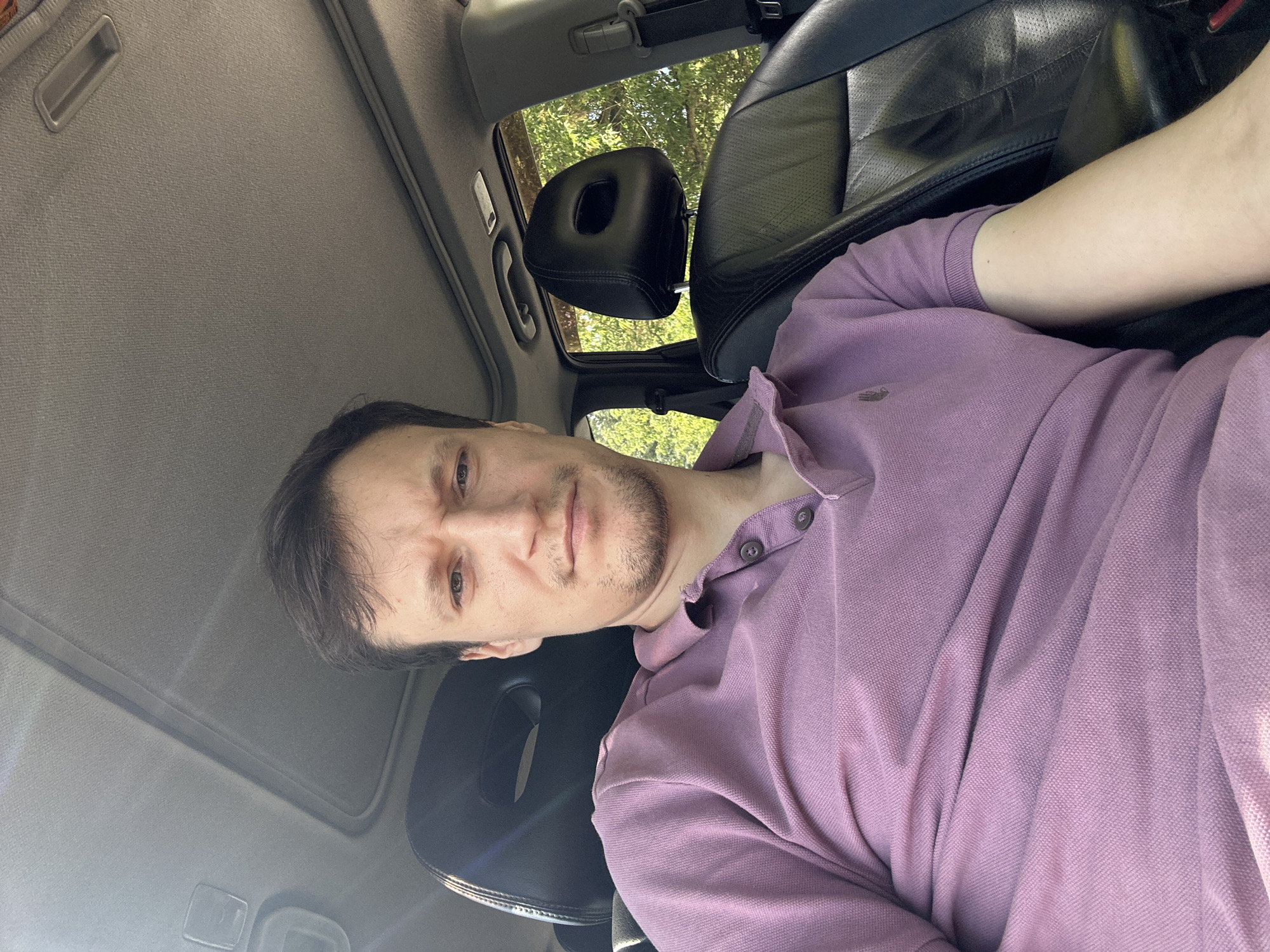 A young man is sitting in the driver's seat of a car. He is wearing a purple polo shirt. The interior of the car is visible, including the headrest of the passenger seat and the car's sunroof. Outside the car, trees are visible, suggesting a natural setting. The image appears to have been taken during the daytime. The exact location in the city of Makiv, Ukraine, is not discernible from the visual information provided.