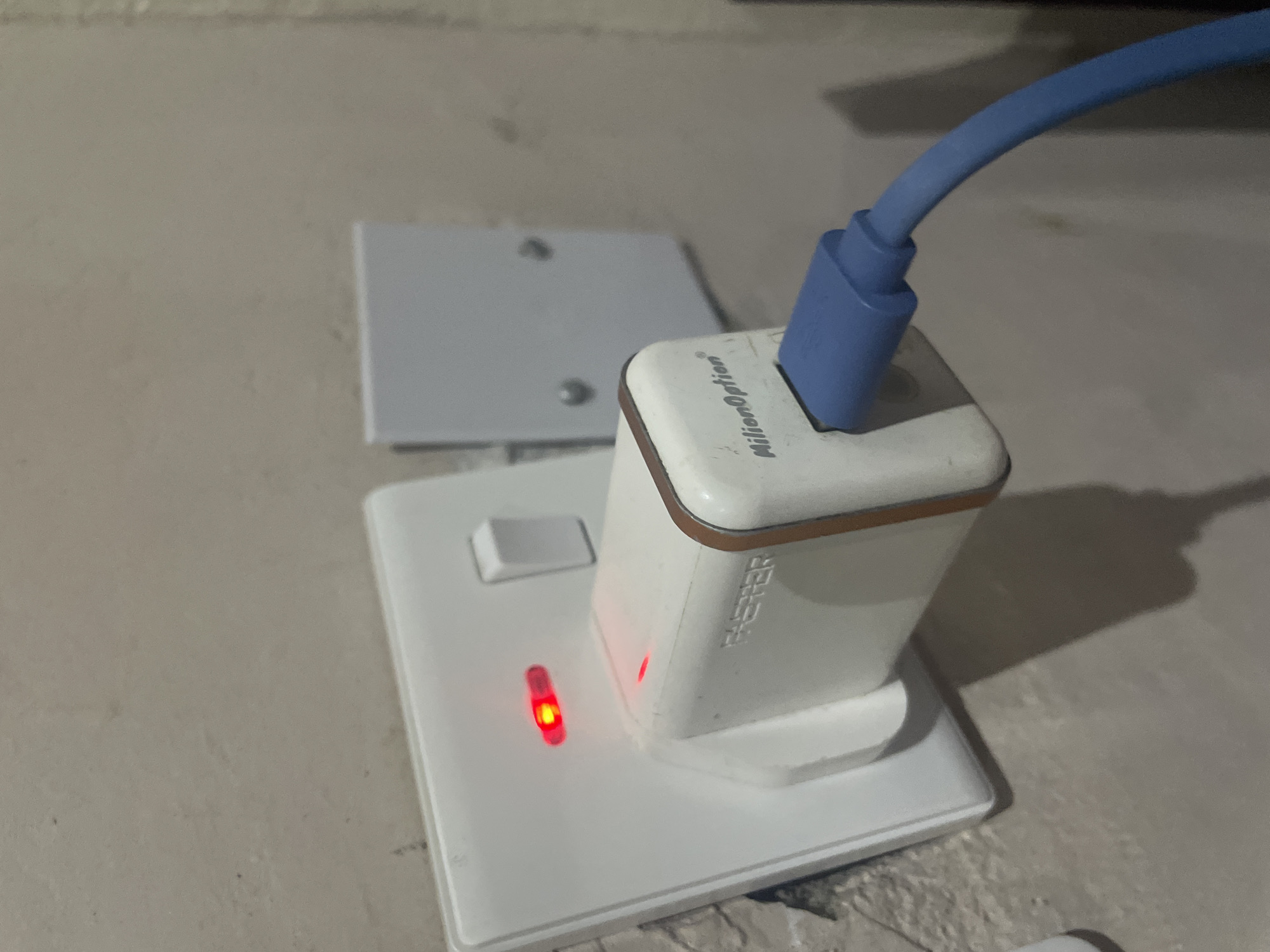 An electrical socket with a red indicator light is mounted on a textured wall. A white power adapter, branded "MillionOption", is plugged into the socket, with a small red indicator light visible on the adapter as well. A blue charging cable is connected to the adapter. To the right of the main socket, a secondary white electrical box is mounted on the wall. The overall scene appears to be an indoor electrical setup. This image is located in Jalingo, Nigeria.
