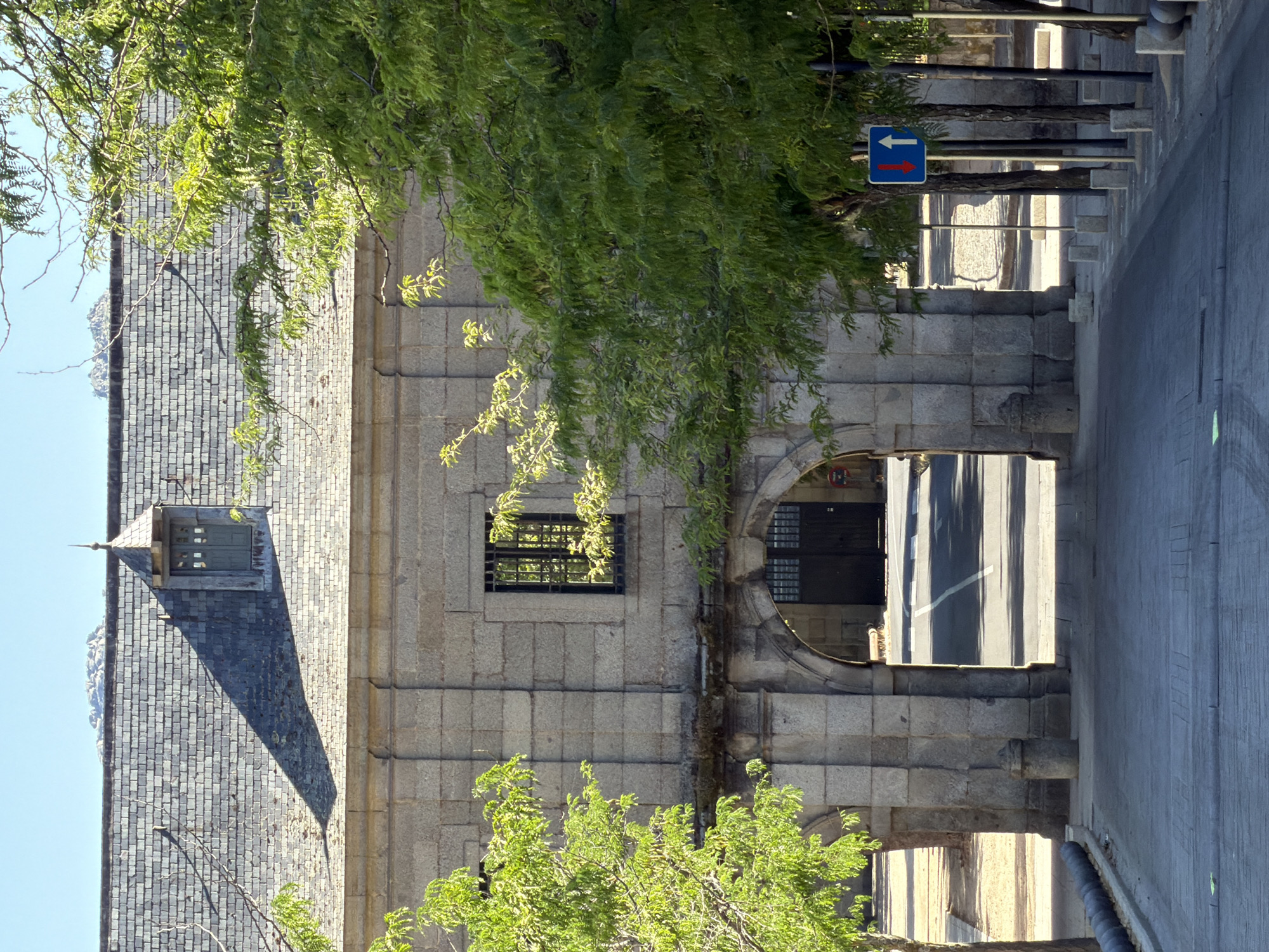 The image depicts the exterior of a stone building with a grey slate roof, likely a historical structure, situated in San Lorenzo de El Escorial, Spain. The building features an arched entryway with heavy wooden doors visible beyond. To the left and right of the entryway, and in front of the building, are trees with vibrant green foliage. A paved pathway leads up to the arched entrance. In the foreground, a grey paved area with some subtle markings is visible. A street with a white line marking the center is seen through the archway. On the right side of the image, there are several trees and a blue traffic sign displaying an upward white arrow and a downward red arrow, indicating two-way traffic. The sky is clear and blue, suggesting a sunny day. The architecture suggests a location with historical significance, possibly related to the Royal Site of San Lorenzo de El Escorial, given the city's association with this landmark.
