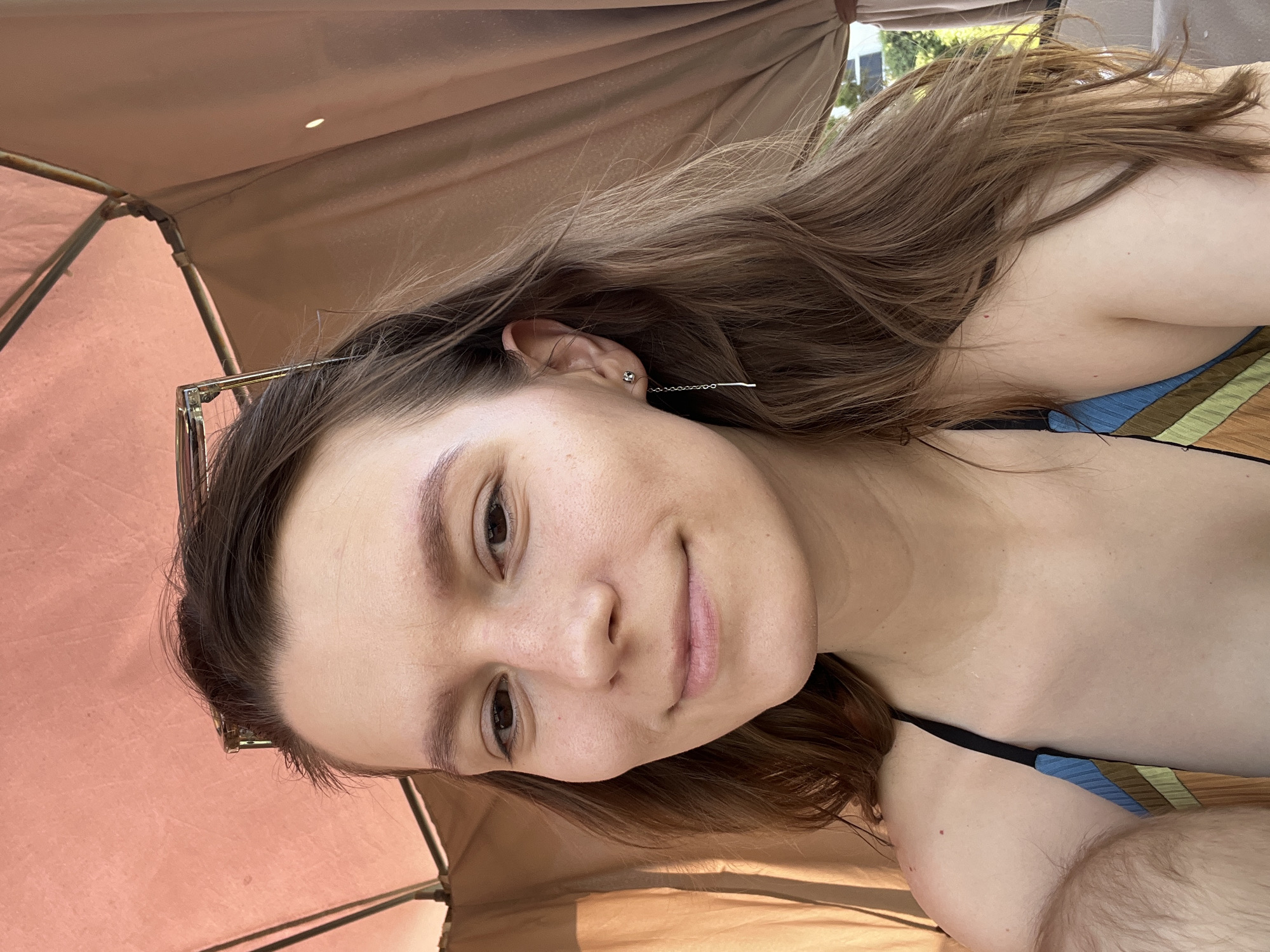The media file is a selfie of a young woman, taken outdoors, likely during the daytime given the bright lighting. She is lying down, possibly under an umbrella or awning, indicated by the textured fabric in the background and the partial view of a frame structure. She has long, light brown hair that is partially obscuring her face and shoulder. She is wearing a bikini top with a striped pattern in blue, brown, and green. A thin, silver chain earring dangles from her ear. The woman has light-colored skin and appears to be smiling softly. Her eyebrows are dark and well-defined, and her eyes are brown. There is a slight tan line visible on her décolletage. The overall impression is casual and relaxed, suggesting a leisure activity like sunbathing. There is no visible text in the media file. The setting might be a beach or poolside given the context of Cyprus and the swimwear.