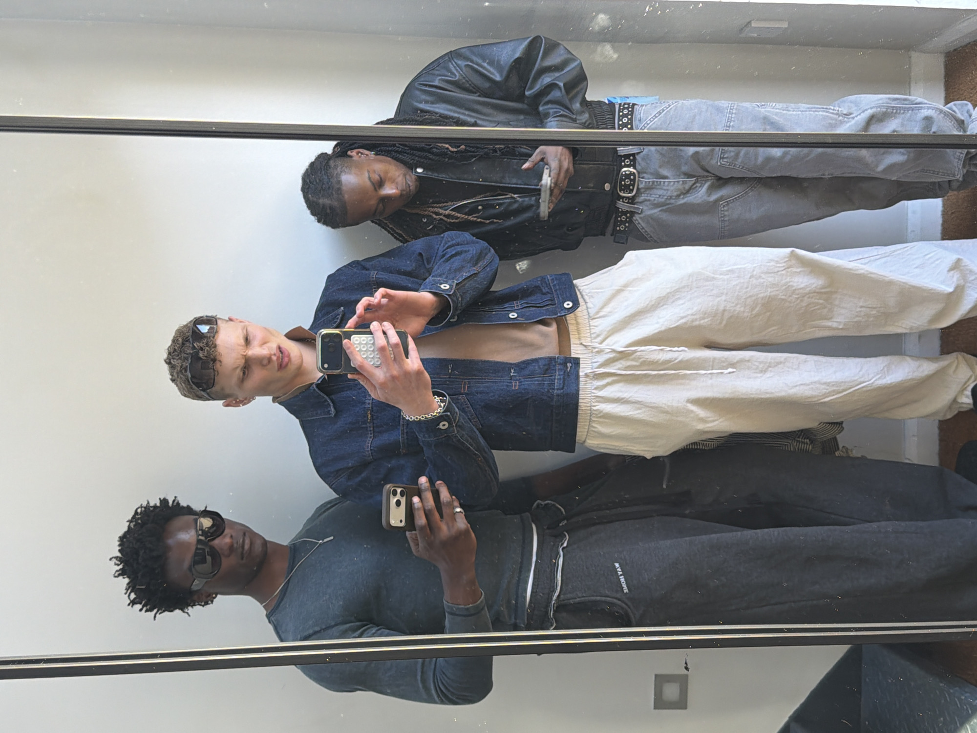 This media file displays a mirror reflection of three young men posing, likely an indoor selfie, in London, United Kingdom. The image is oriented with the subjects lying horizontally across the frame.
In the upper part of the reflection, a dark-skinned man with long, dark braided hair is visible. He wears a black leather jacket over a dark top and light grey cargo pants, secured with a prominent black studded belt. He is looking downwards and holding a phone in his left hand, which is partially obscured.
Centered in the reflection is a light-skinned man with short, curly light brown hair, his sunglasses pushed up onto his head. He is dressed in a blue denim jacket over a light-colored top and light beige or off-white pants. He holds a phone with both hands, appearing to interact with it, and displays a slightly squinted or winking expression. A silver chain bracelet is visible on his left wrist.
In the lower part of the reflection, a dark-skinned man with short dark curly hair is seen. He wears dark sunglasses, a dark long-sleeved top, and dark sweatpants. He holds a phone in his right hand. Mirrored text, "EOPH AVM" (likely "MVA HOPE" when unmirrored), is visible on the upper thigh of his sweatpants.
The background behind the reflected subjects is a plain, light-colored wall. A thin black frame delineates the mirror from its surroundings. A dark floor is visible at the very bottom edge of the reflection. The overall scene captures a casual group mirror shot, highlighting different streetwear styles among the young men.