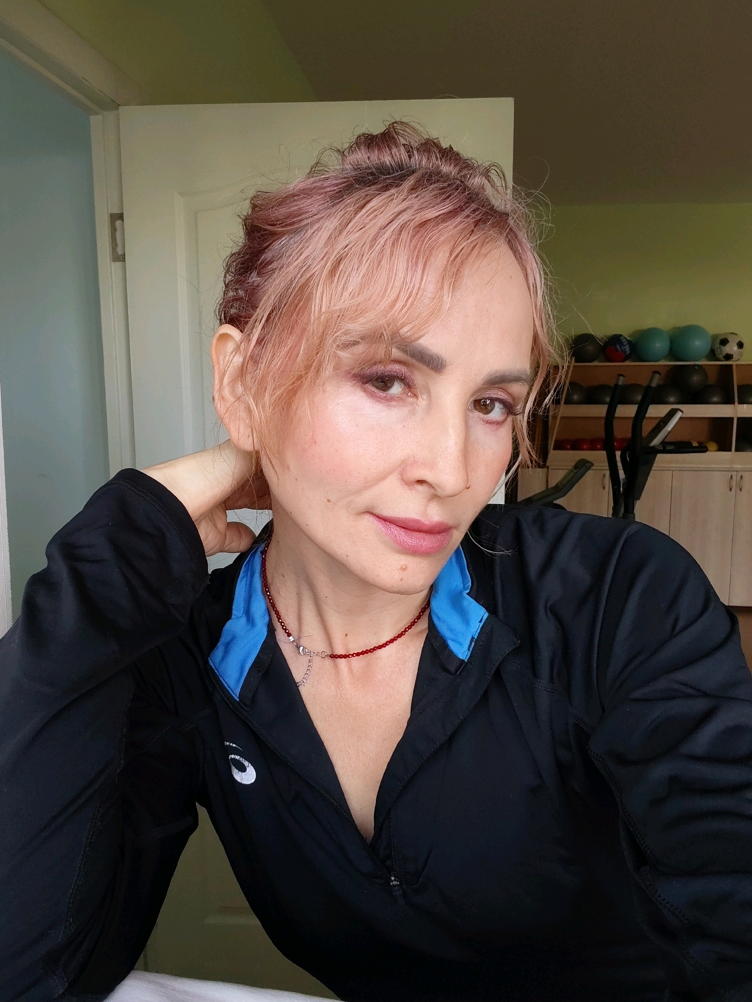 A medium-shot, eye-level, horizontal photograph shows a woman with light skin and auburn hair styled in a bun. She is wearing a black athletic jacket with a blue collar and a red beaded necklace. Her right arm is bent behind her head, and her left hand is resting on her neck. The woman is looking towards the camera, with her head slightly tilted.
The background consists of a room with a light green wall. Shelves with exercise equipment, including balls and what appears to be an elliptical machine, are visible. A white door is on the left side of the image.
The image does not provide any specific information or clues that would allow for the identification of its location in the city of Chaikovsky, Russia.