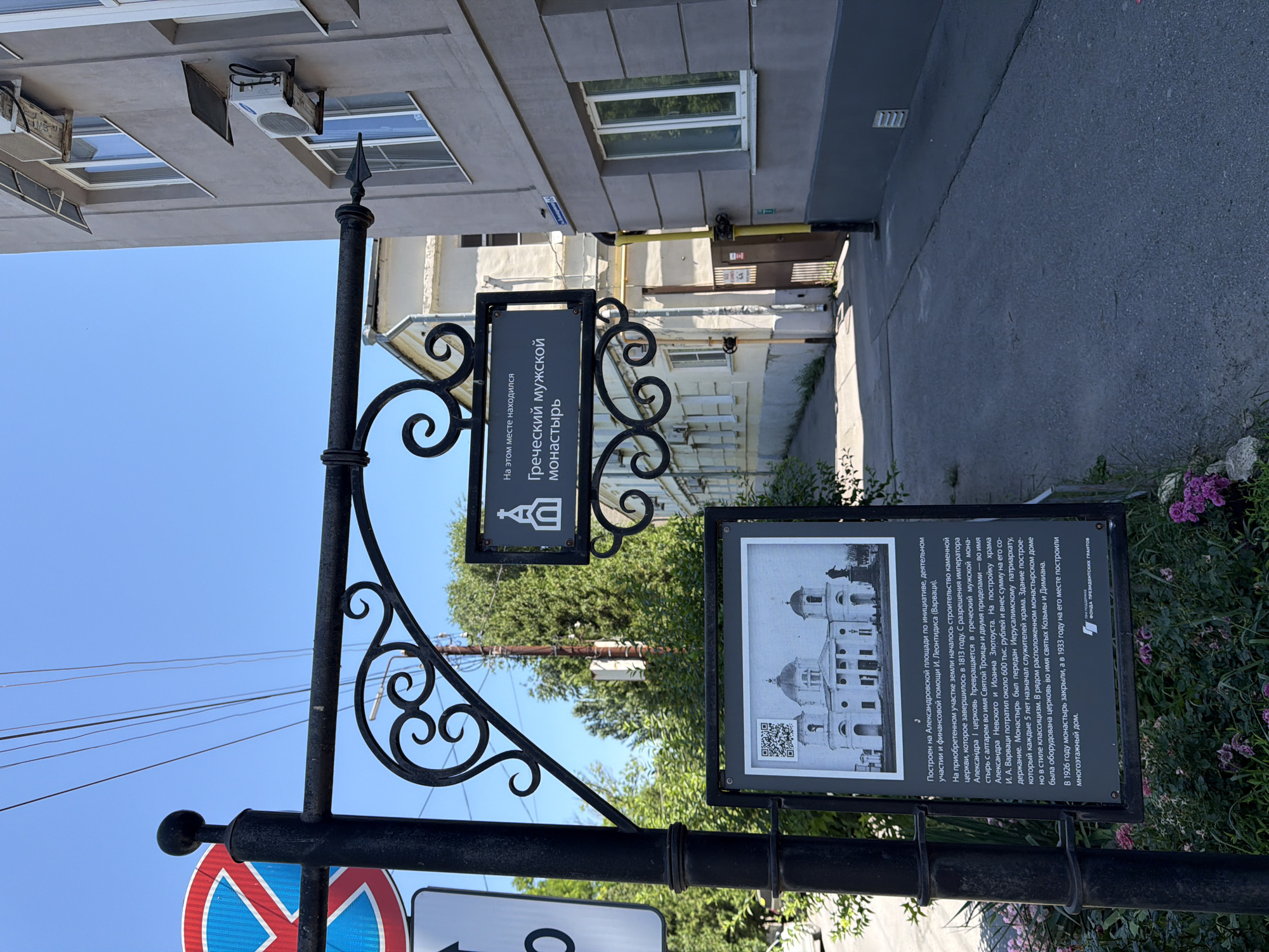 This is a photograph taken in Taganrog, Russia, depicting a street scene with informational signs. The image is oriented vertically. In the foreground, a black wrought-iron pole holds two signs. The upper sign, also black, has white text in Cyrillic that translates to "Greek Male Monastery was located here" and displays an icon of a church. Below it, a larger black sign has a black and white illustration of a church and a QR code. The text on this larger sign, also in Cyrillic, provides a historical description of the monastery's founding in 1813, its patrons, architectural style, and eventual closure and demolition in the 1920s and 1930s. A red and blue traffic sign is also visible on the pole.
In the background, a modern building with air conditioning units is visible, suggesting an urban setting. Sunlight indicates it is daytime, with clear blue skies. The overall scene captures a blend of historical information and contemporary urban life, marking a significant historical site within the city.
