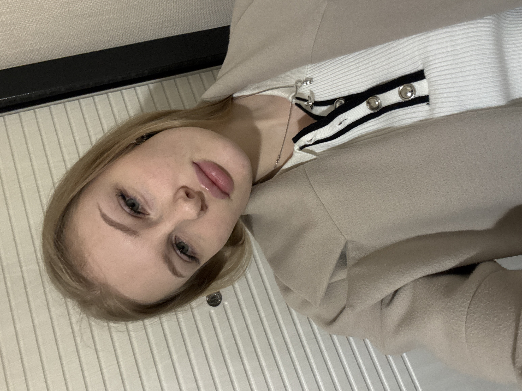 A fair-skinned woman with blonde hair is lying on a horizontal surface. She is wearing a beige jacket over a white sweater with black and white stripes and silver buttons. A silver necklace is visible around her neck. The background features white, vertically-aligned panels. The location is presumed to be in Perm, Russia.