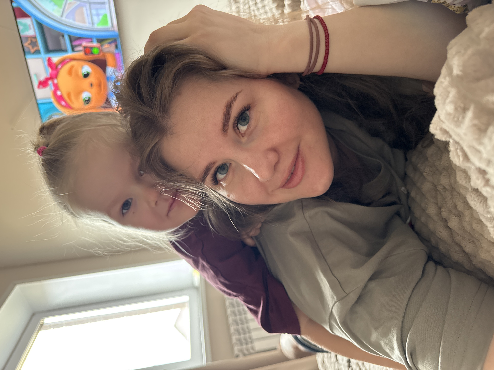 The image shows a woman and a young child, likely a mother and daughter, lying down indoors. The woman is in the foreground, looking at the camera with a slight smile. She has light skin, brown hair, and light blue eyes. On her wrist is a thin red bracelet. Behind her, partially obscured, is the child, with blonde hair and blue eyes. A TV screen is visible in the background, displaying a cartoon character. A window provides natural light into the room. The location could be Gornyi, Russia based on the prompt. They appear to be in a domestic environment, likely a bedroom.
