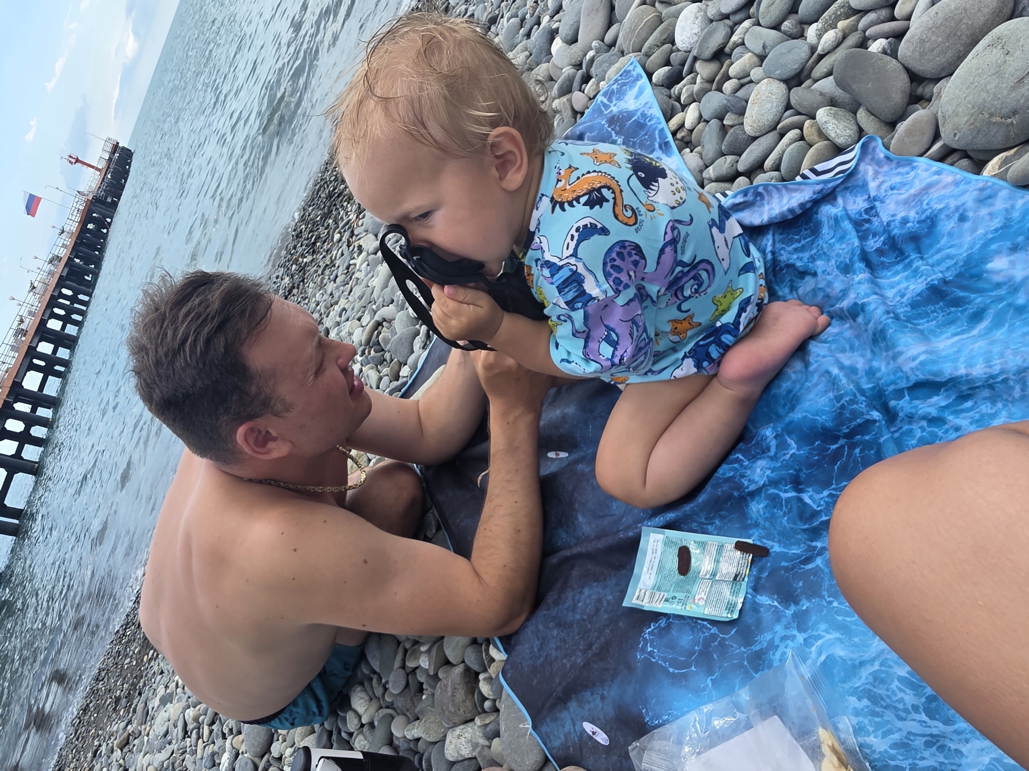 A man and a young child are on a rocky beach in Sochi, Russia. The man is kneeling on a blue towel with a wave pattern and is looking at the child. The child is sitting on the towel and holding a black object, possibly a mask, up to their face. The child is wearing a light blue rash guard with an octopus and seahorse pattern. In the background, there is a pier with a Russian flag and a lighthouse. The sea is visible behind the pier. A package of chocolate and a plastic bag with some food items are on the towel next to the child. The weather appears to be overcast.