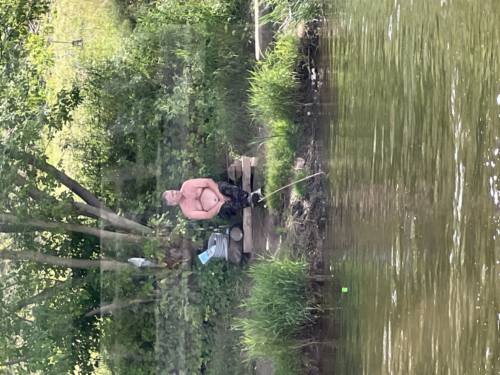 A middle-aged man with graying hair and a bare torso is sitting on a wooden platform near the bank of a river. He is wearing camouflage-patterned pants and dark shoes. Next to him on the platform is a gray bucket with a blue object inside. A fishing rod is propped up against the platform. The river is calm and appears greenish in color. The background consists of lush green trees and vegetation. The scene is set in a natural environment, possibly a park or a recreational area.