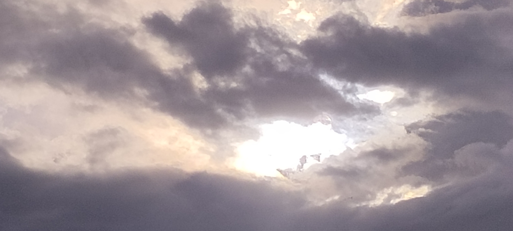 The image shows a cloudy sky. There are no visible features that indicate the location in Lhokseumawe, Indonesia. The sky is filled with a mix of dark gray and lighter, sunlit clouds. The sun is partially obscured by the clouds, creating a bright patch of light in the center.