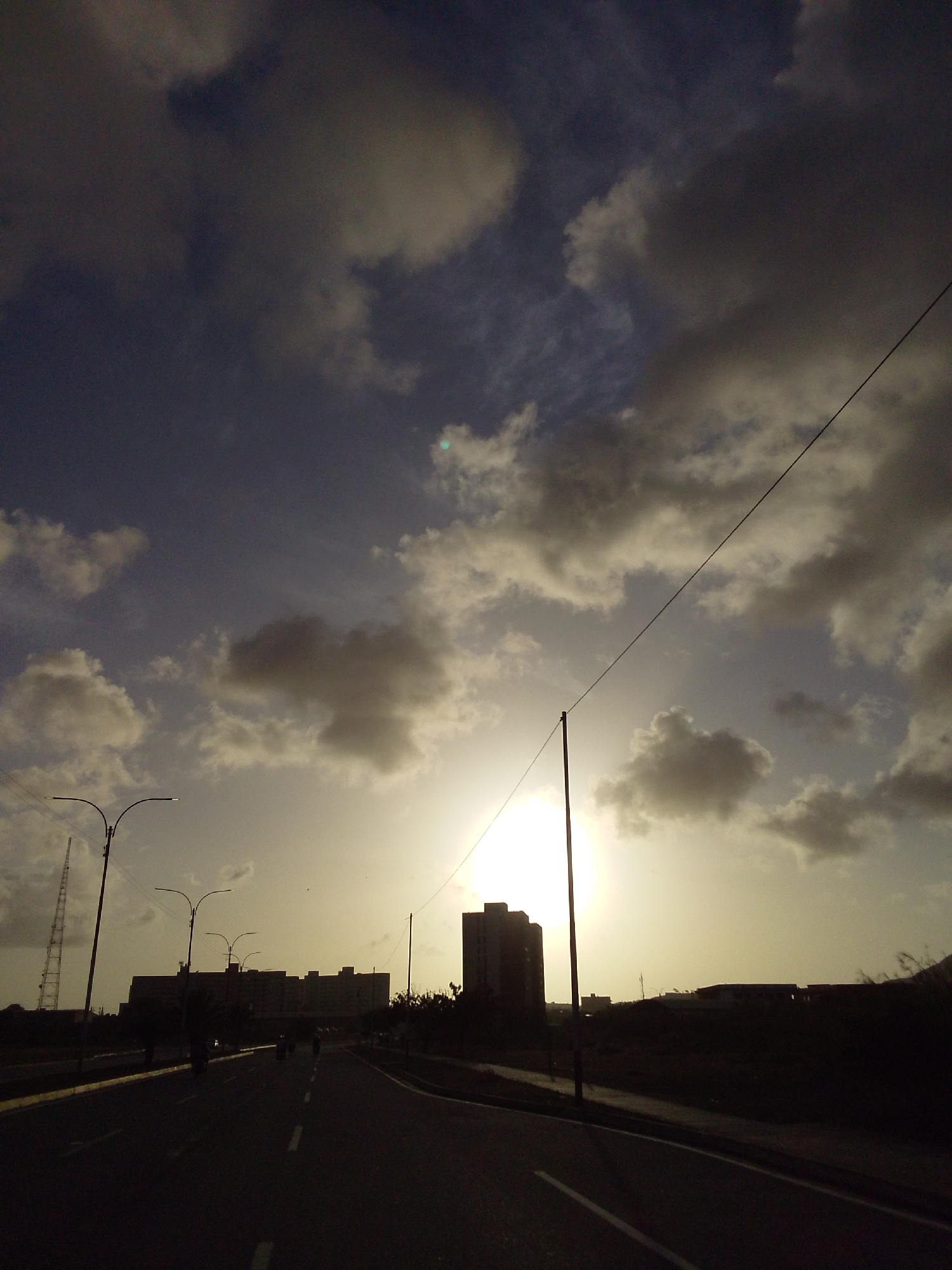 An eye-level, wide shot shows a street scene at dusk in an urban environment. The sky is filled with dramatic, heavy clouds with the sun peeking through, creating a bright, hazy glow. A thin, dark power line stretches diagonally across the upper portion of the frame. In the mid-ground, a dark, silhouetted cityscape is visible with several multi-story buildings, including a taller, blocky building on the right. A few streetlights with curved poles line the left side of the road, and a tall, lattice tower is visible in the far left background. The foreground is a dark asphalt road with faint white lines marking the lanes. Several motorcycles are moving along the road, their silhouettes dark against the lighter background. The overall mood is atmospheric and slightly ominous due to the dramatic sky and silhouettes.