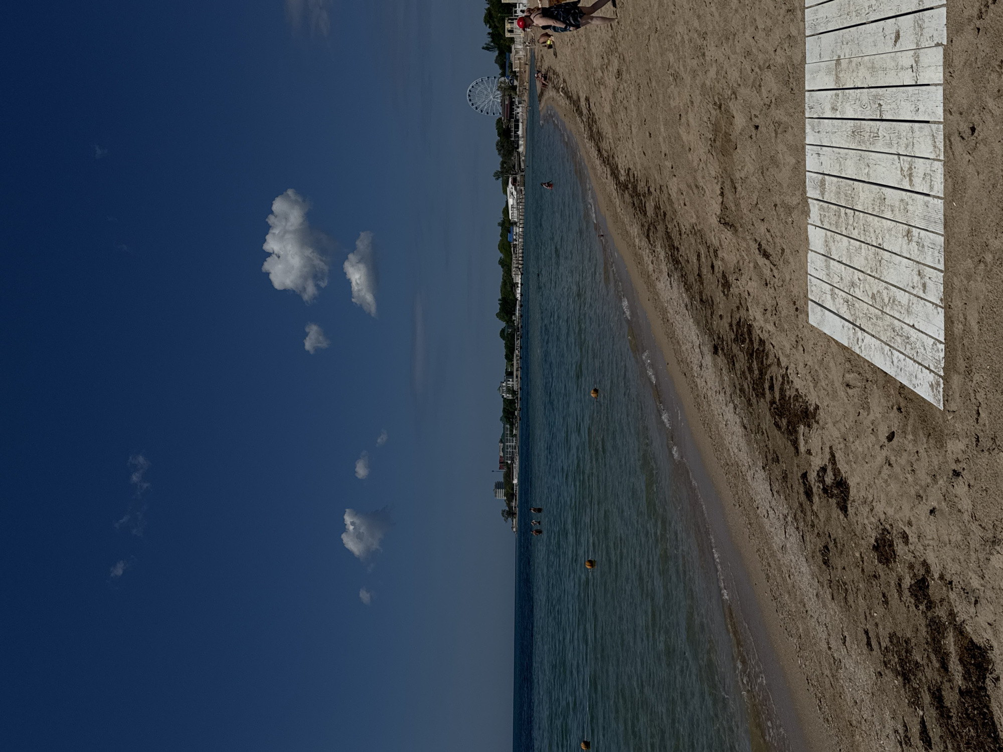 The image shows a beach scene in Yevpatoriya, Ukraine. The composition is dominated by a stretch of sand, transitioning from a wooden walkway in the foreground to a more natural, seaweed-strewn section closer to the waterline. The water is calm with some buoys floating in it. In the distance, there are buildings along the coast and a Ferris wheel is visible. The sky is blue with scattered clouds. There are a few people walking near the Ferris wheel.
