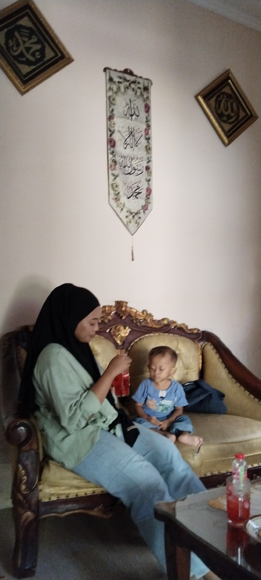 In Peganjaran, Indonesia, a woman wearing a black hijab and a light green tunic sits on a gold-accented couch. She is holding a red drink in a clear bottle. Beside her sits a young boy wearing a blue t-shirt and pants, with his eyes closed. The couch is ornate, with dark wood trim and gold detailing. On the wall behind them hangs a long, rectangular banner with Islamic calligraphy and floral designs, flanked by two framed Islamic calligraphy pieces. A dark wooden coffee table is in the foreground, with another red drink in a bottle on it. The environment appears to be an indoor setting.