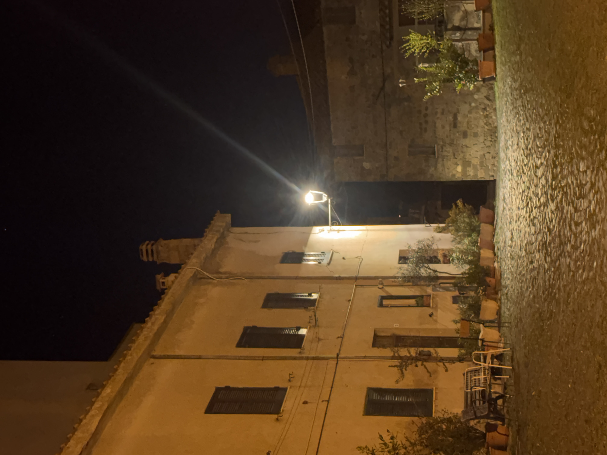 The image depicts a night scene in Castelnuovo di Porto, Italy. The view is angled upwards towards a multi-story building with closed window shutters. The building is illuminated by a street lamp. Adjacent to the building is a stone wall. A cobblestone pathway runs alongside the wall. Potted plants are situated along the base of the building and wall. There are some chairs and a table on the right side of the image. The sky is dark.
