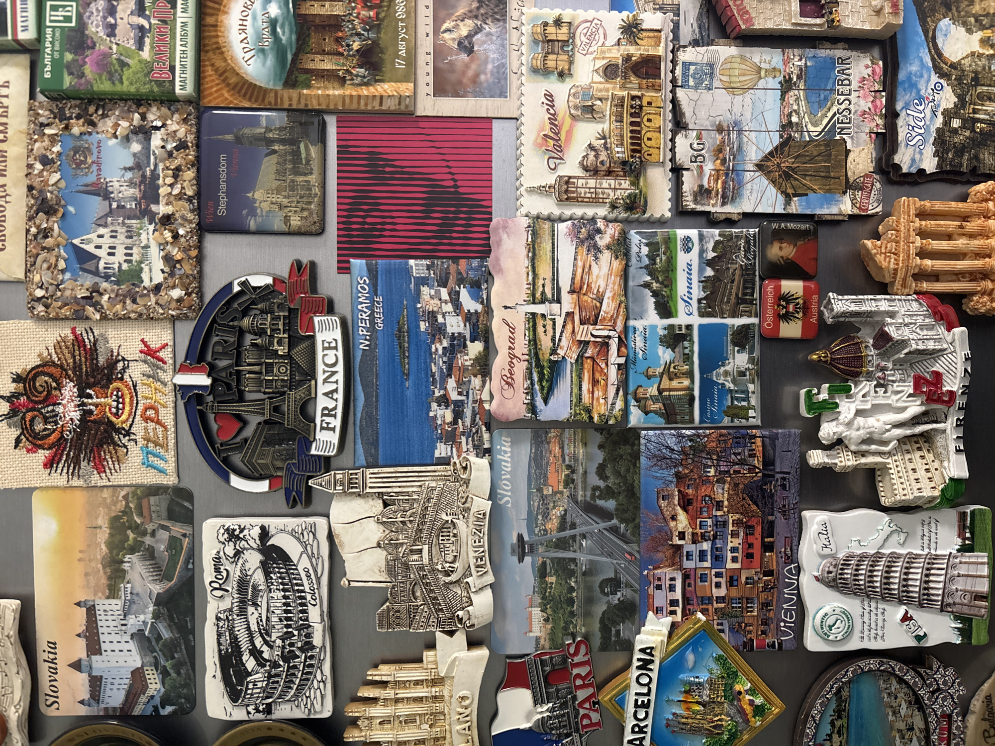 The image is a close-up shot of a collection of souvenir magnets arranged on a gray surface. The magnets are from various cities and countries around the world, including Barcelona, Paris, Venice, Vienna, Slovakia, Rome, Greece (N. Peramos), Valencia, Nessebar, Side, and Bulgaria, among others. Some magnets display city skylines, famous monuments (like the Colosseum in Rome and the Leaning Tower of Pisa), landscapes, and other images. There are also magnets with the names of the places and flags. Some magnets have 3D elements or textures, and overall, the arrangement suggests a collection of travel memories.