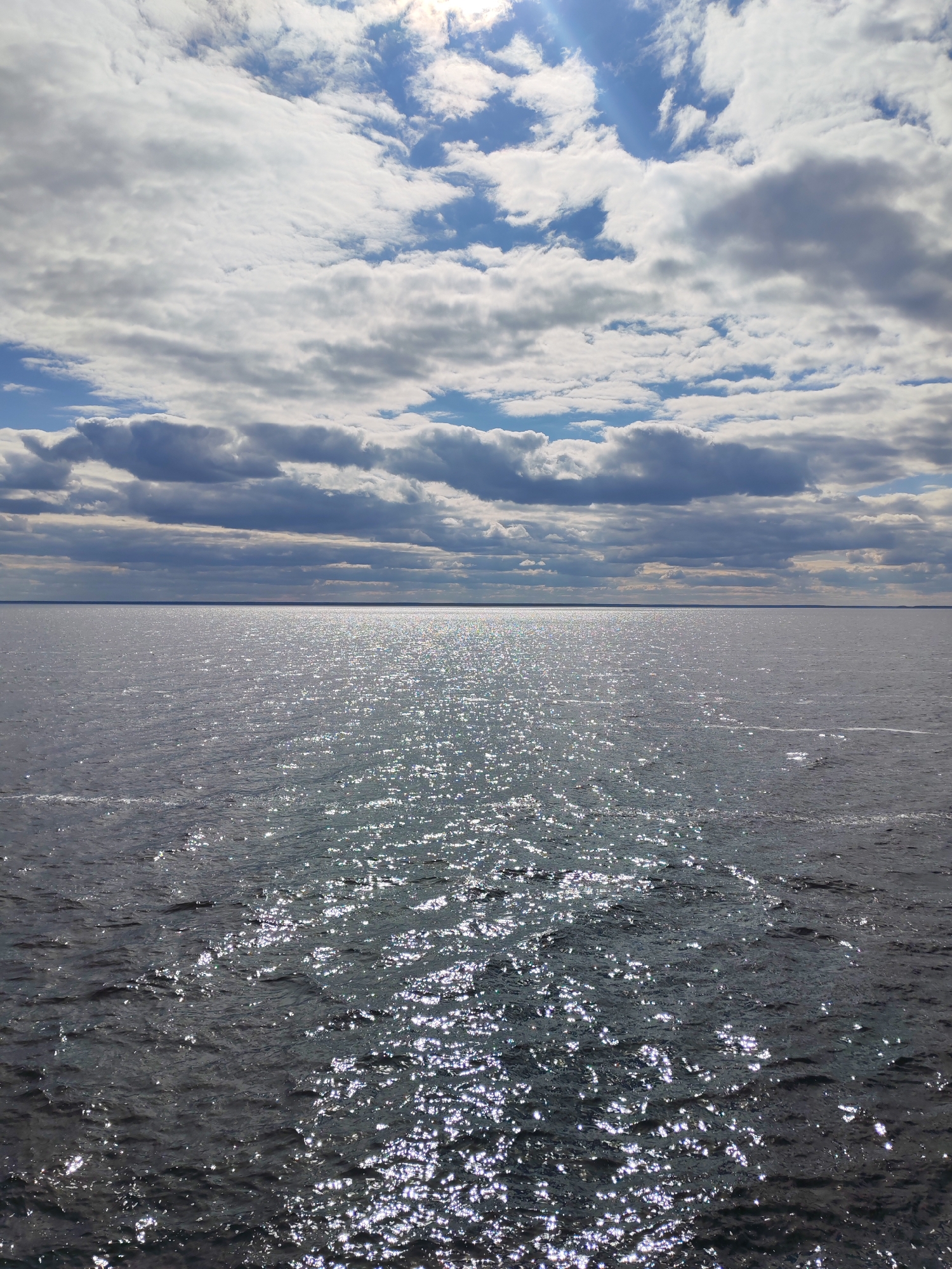 The image shows a body of water under a partly cloudy sky. The water reflects sunlight, creating a shimmering effect. The horizon line is visible in the distance. The location is likely in the vicinity of Vasil'evskoe, Russia, given the implied geotagging. There are no people, subjects, objects, actions or interactions that can be gleaned from this image, other than the presence of water and the natural environment.