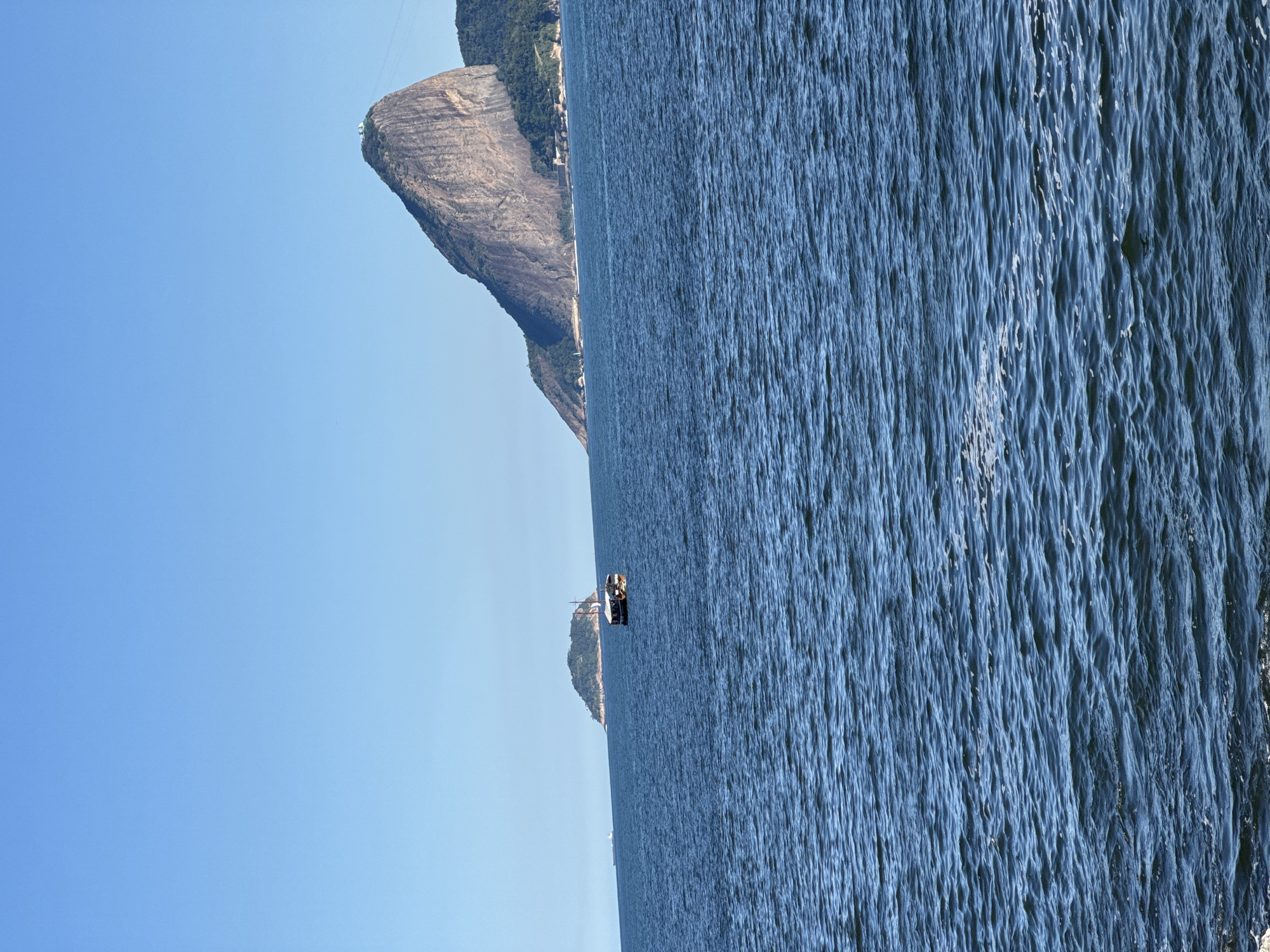The image shows a wide, blue ocean under a clear, light blue sky. In the distance, a large, rocky mountain with a green top rises from the water. There is a small island covered in trees to the left of the mountain. A single, small wooden boat with a dark roof is sailing on the ocean. The water is slightly choppy with small waves. The scene is likely from Rio de Janeiro, Brazil, given the iconic mountain which is Sugarloaf Mountain.