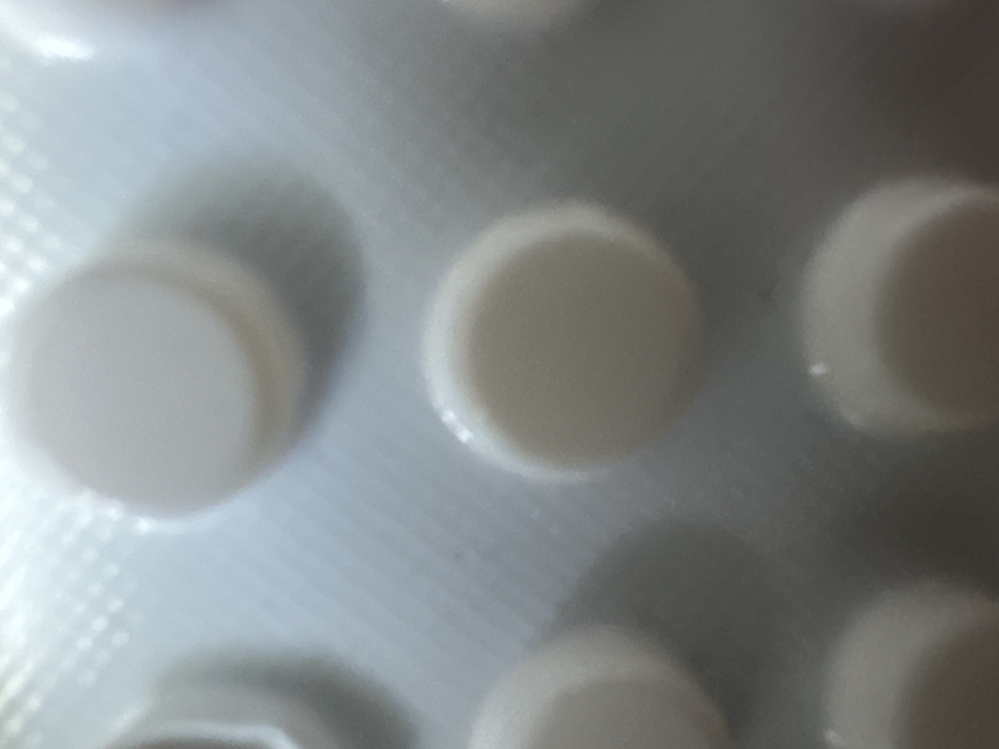 The image displays a close-up, out-of-focus view of a blister pack containing several round, white pills. The pills are arranged in a grid pattern, with some in focus and others blurred. The packaging material is a light gray or silver color, with a slightly textured surface that creates a pattern of small squares or dots when viewed closely. The lighting appears to be artificial and is reflecting off the glossy surface of the packaging and the pills, creating bright highlights. There is no visible text in the image. The overall impression is one of a medical product, likely medication, presented in its packaging. The focus is on the pills and their container, with no other elements visible to suggest a specific time of day, weather, location, or emotional context beyond the functional purpose of the object. The scene is a still life, focusing solely on the pharmaceutical product.