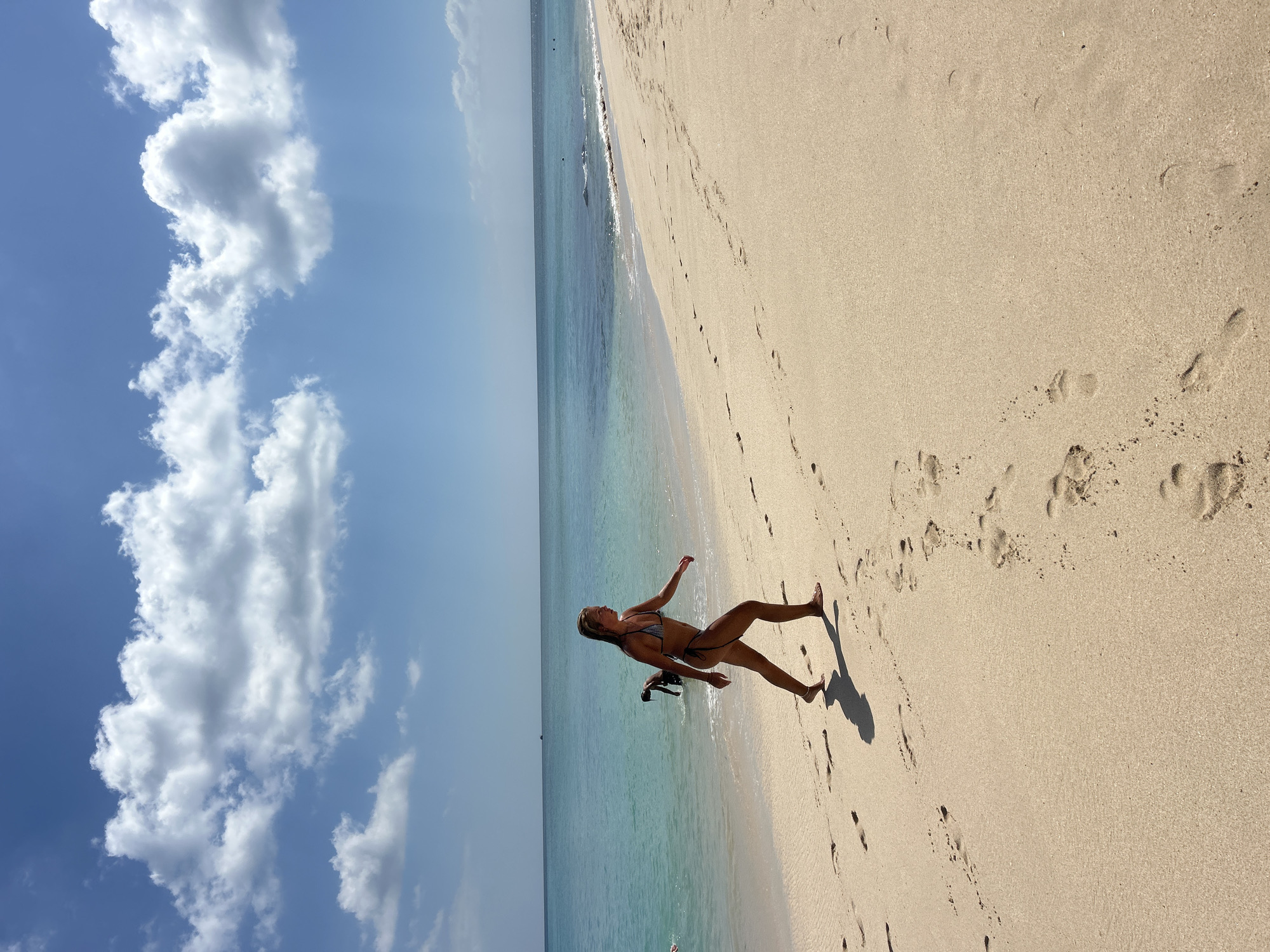 A young woman in a bikini walks along a sandy beach with clear turquoise water and a bright blue sky with white clouds. The woman is walking from left to right in the frame, with her shadow cast on the sand. There are footprints in the sand leading away from her. In the background, a person is partially submerged in the water. The scene is likely a tropical beach location.