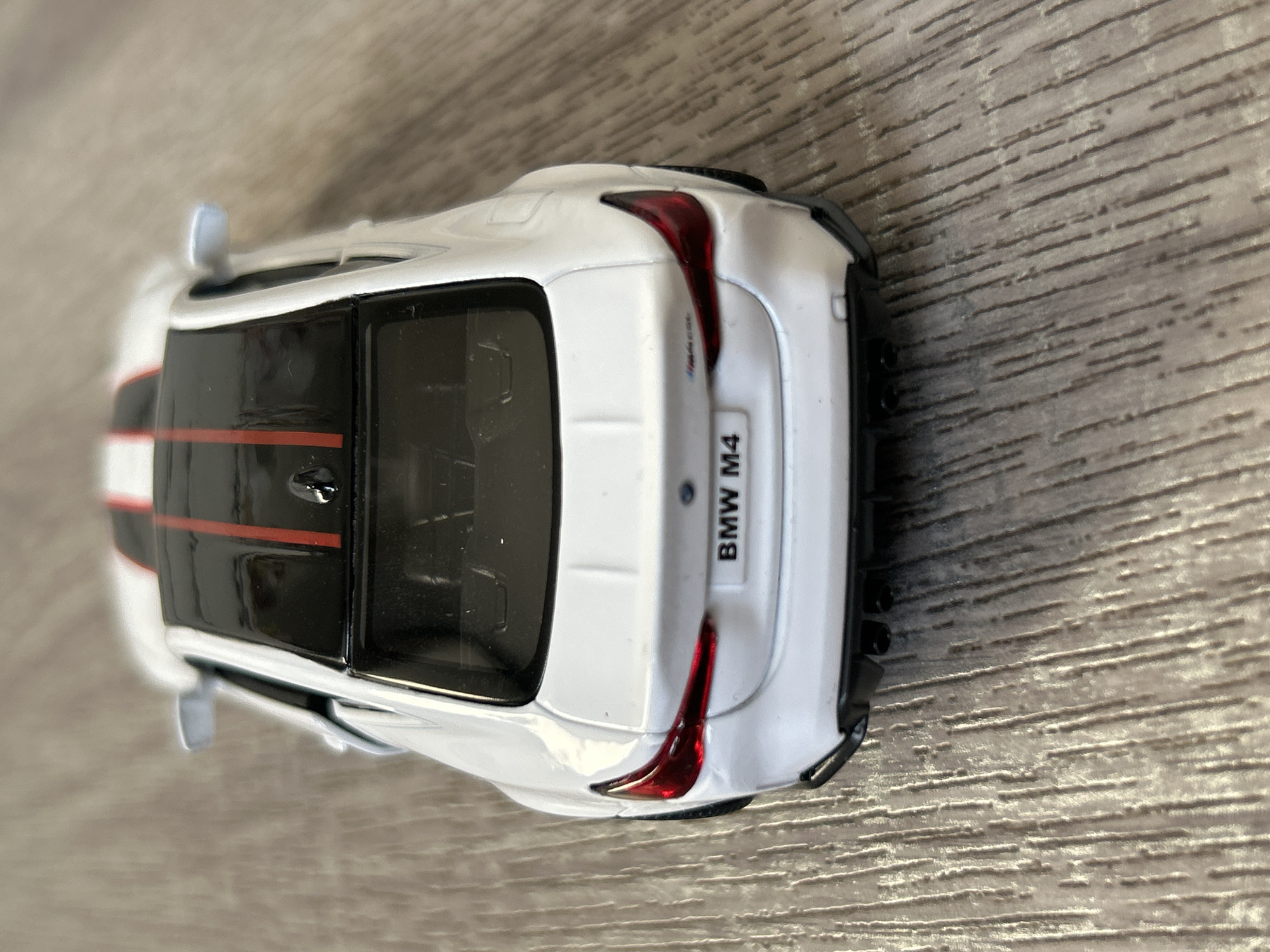The image shows a close-up, top-down view of a white toy BMW M4 car resting on a wood-like surface. The car has a black roof with two red stripes. The taillights are red, and the license plate reads "BMW M4". The setting appears to be indoors due to the controlled lighting and background surface. There is no information to conclude that the location is in the city of Strasbourg, France.