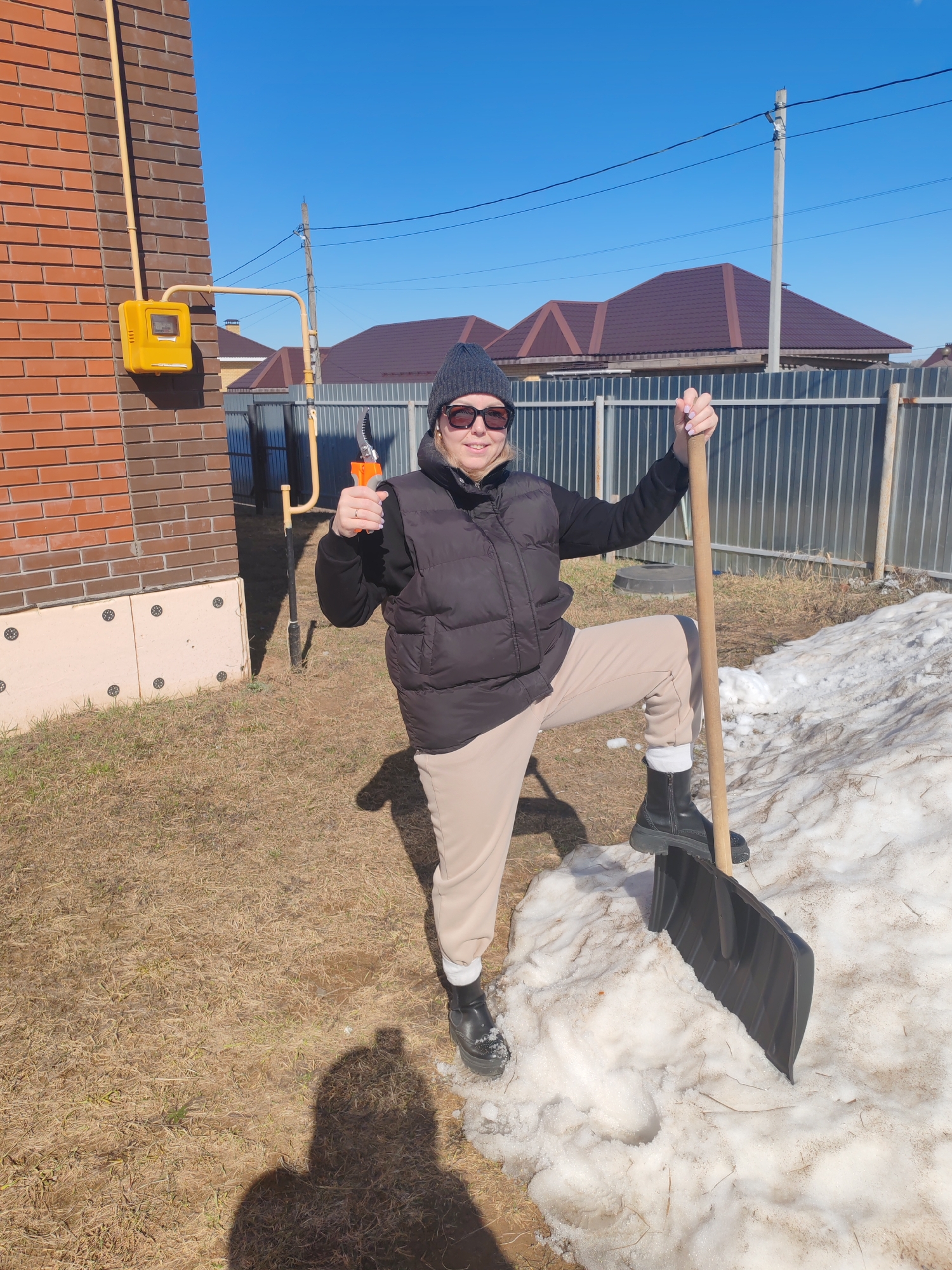 The image shows a woman standing in a yard in Chaikovsky, Russia. She is wearing a dark grey knit hat, sunglasses, a black puffy vest over a dark sweatshirt, light tan pants, and black boots. She has a pair of orange and silver pruning shears in her right hand, giving a thumbs-up gesture. Her left hand is holding the wooden handle of a large black shovel, with her right foot resting on the shovel's blade which is stuck into a pile of snow.
Behind her is a brick building with a yellow gas meter attached to its side. A metal fence runs along the background, behind which are houses with brown roofs. The sky is clear and blue. The ground is covered with dry grass and patches of snow. Shadows are cast by the sunlight.