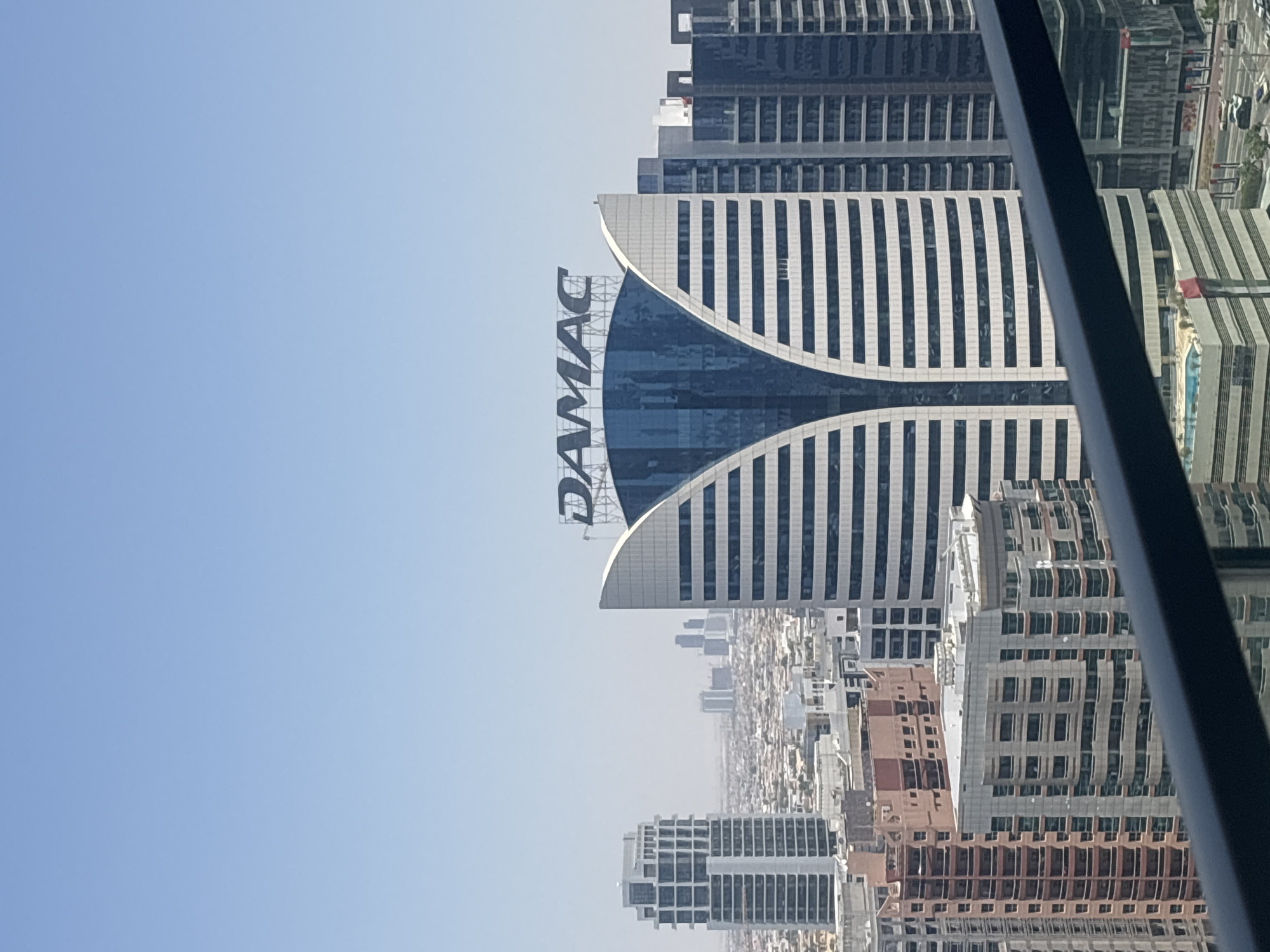 The image shows a cityscape in Dubai, United Arab Emirates, on a clear, sunny day. The foreground features a dark railing, suggesting the photo was taken from a high vantage point, possibly a balcony or rooftop.
Dominating the mid-ground is a distinctively shaped building with the prominent "DAMAC" logo at its peak. This building features a curved facade with alternating horizontal bands of white and blue, creating a striped pattern. The blue sections appear to be glass, reflecting the sky and surrounding buildings.
To the left of the DAMAC building, another tall, rectangular skyscraper with a white facade and a grid-like pattern of windows stands out. In the background, a cluster of other high-rise buildings of varying architectural styles can be seen, suggesting a dense urban environment. The overall impression is of a modern metropolis with a focus on contemporary architecture. The sky is a pale blue, with no visible clouds, and a slight haze is present, softening the distant view of the city. The scene is devoid of people or any discernible activity.