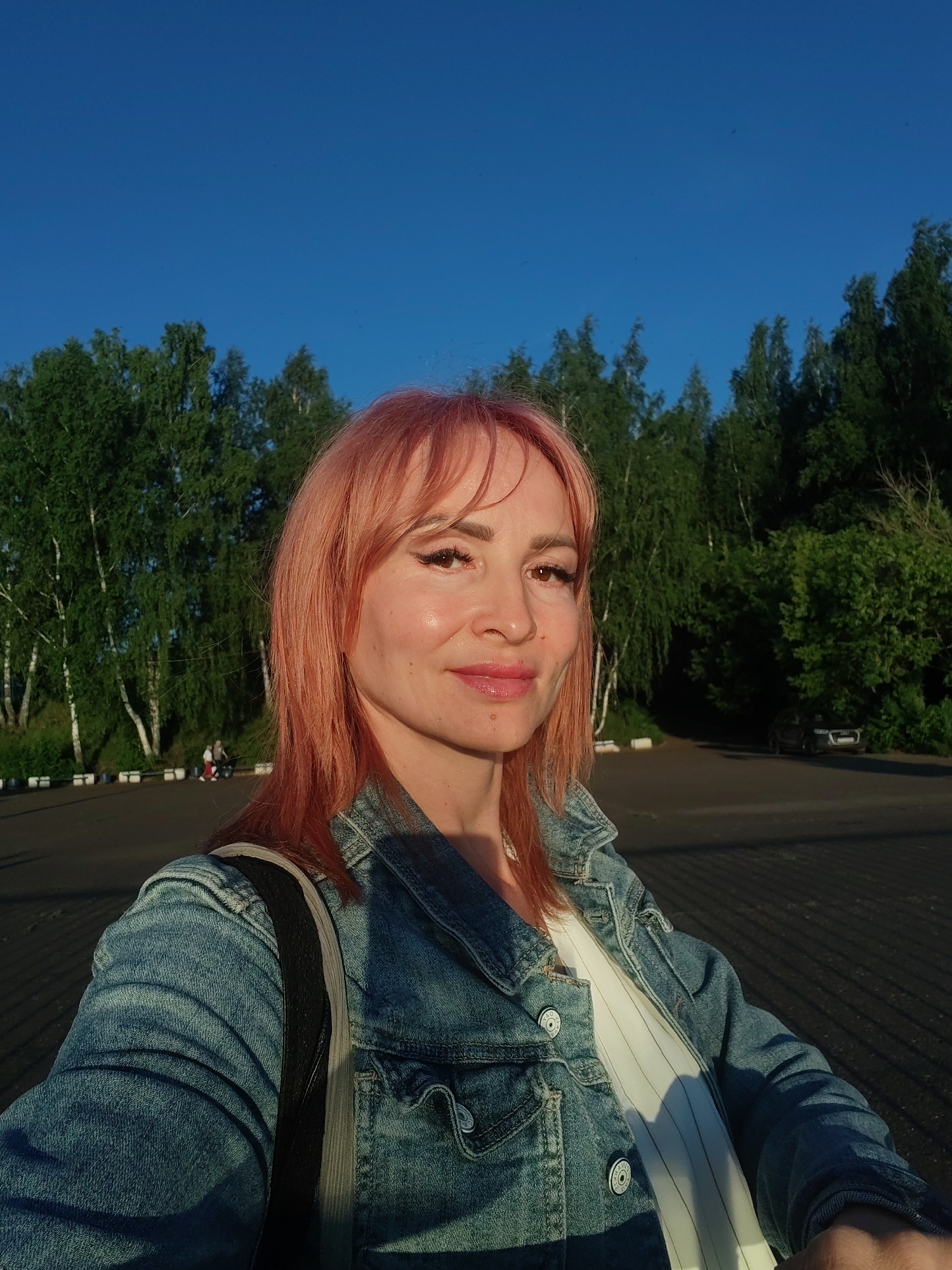 A woman with pink hair and a denim jacket stands in front of a line of trees under a clear blue sky. She is taking a selfie, and her face is lit by the sun. In the background, there are more trees, a paved area, and a few cars parked. The image appears to be taken in the city of Chaikovsky, Russia.
