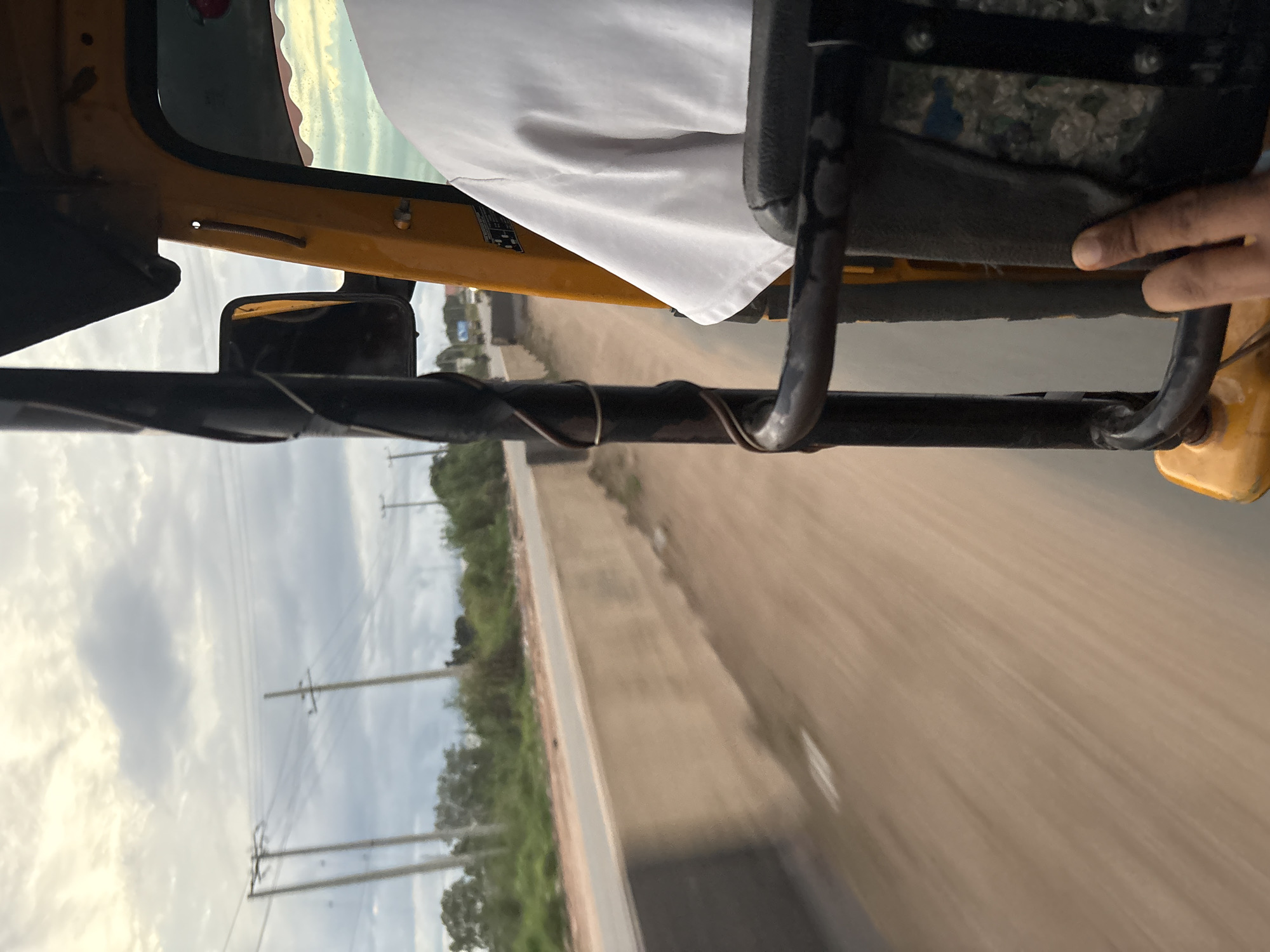An overhead shot captures a person's hand and arm from the elbow down, with their index finger extended and touching a dark grey textured seat. The hand and arm are positioned in the lower right quadrant of the frame. The person is inside a yellow vehicle, likely a rickshaw or auto, with a portion of their white t-shirt visible. The vehicle's interior features black metal framing and a dark, padded seat. The exterior view through the vehicle's open side shows a blurred landscape of a dirt road or path running alongside a concrete barrier, with greenery and utility poles in the background under a cloudy sky. This scene is indicative of travel within Onitsha, Nigeria, a bustling commercial hub known for its vibrant street life and transportation methods like auto-rickshaws.