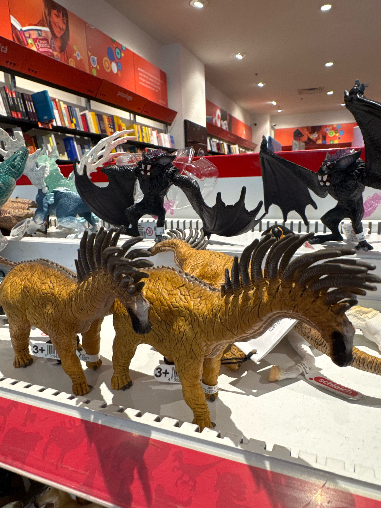 This is a medium shot, eye-level, indoor, brightly lit photograph taken at a bookstore in Carini, Italy. The focus is on several toy figurines displayed on white shelving with a red decorative border along the bottom. In the foreground, two toy horses with manes of stiff, dark brown spikes and textured, golden-brown bodies are positioned closely together. The horse on the left has a tag reading "3+ 4 05943", and the horse on the right has a similar tag. Behind the horses, there are two black, bat-like toy creatures with leathery wings, sharp teeth, and red eyes. One of these bat figures is positioned to the right, and another is to the left and slightly behind. In the background, there are shelves stocked with books of various colors. The upper part of the image shows a large, orange poster with a child reading a book, and other colorful circular graphics. The overall scene suggests a children's section or display within a bookstore.