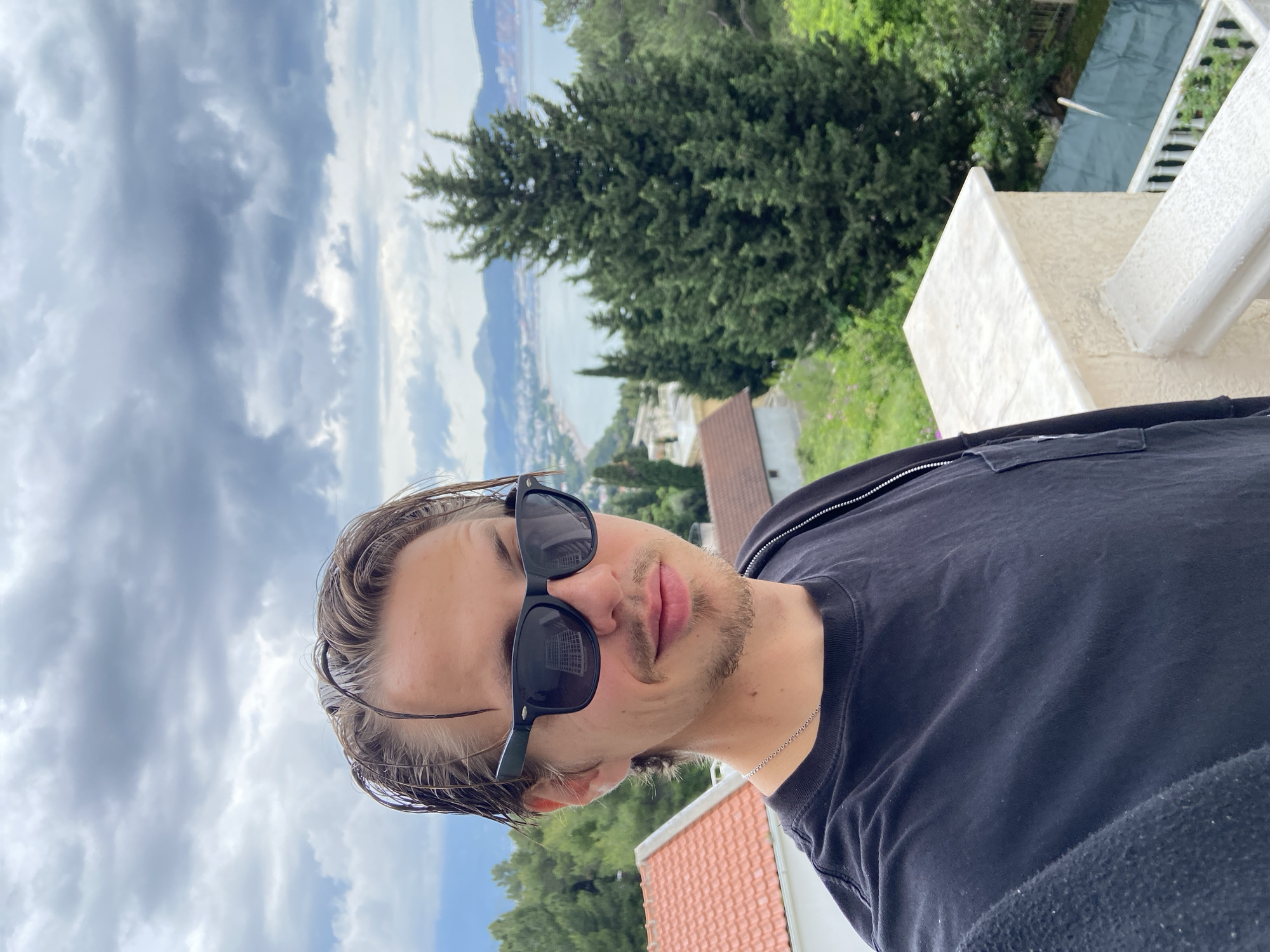The image shows a man wearing sunglasses and a black shirt. He is standing outdoors, possibly on a balcony or rooftop, with a view of the bay visible in the background. In the background there are houses with red roofs and also trees. There are dark clouds. The exact location appears to be Šušanj, Montenegro.