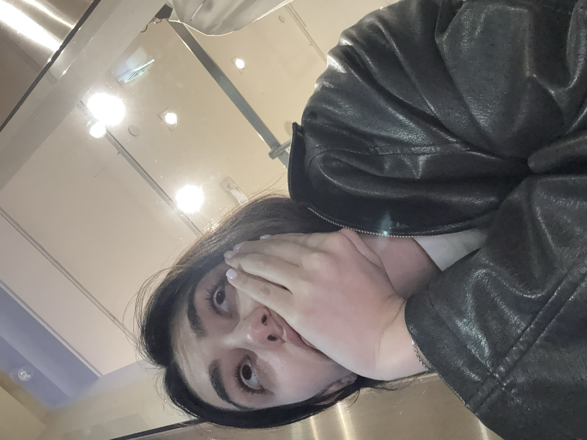 A young woman with dark hair and large brown eyes poses for a selfie with her hand partially covering her face. She wears a black leather jacket. The reflection in the glossy surface reveals a bright, modern interior with overhead lighting, indicating a contemporary indoor space. The environment isn't detailed enough to determine the specific location in Milan.