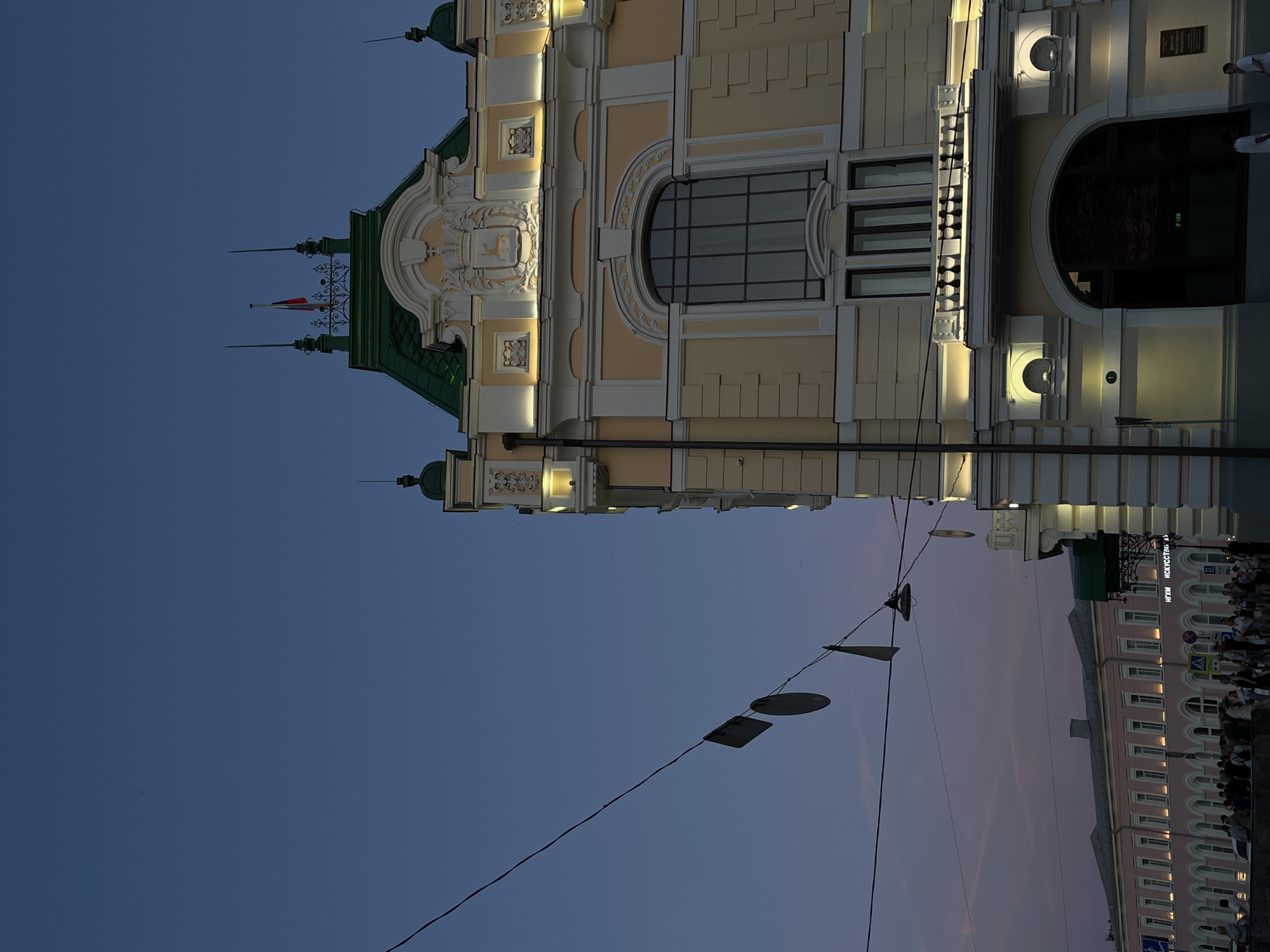 This image captures a dusk view of the Nizhny Novgorod State Art Museum, located at Kremlin, building 5, Nizhny Novgorod, Russia. The ornate, yellow facade of the museum is illuminated by warm lights, highlighting its architectural details. A prominent green roof with spires and a flag adorns the top of the building. In the foreground, wires strung across the sky are adorned with several flat, square and circular objects, possibly decorations or signage. Below, a long, pink building with numerous arched windows and lights stretches along the street, where a crowd of people is gathered. The sky is a deep blue with subtle pink hues near the horizon, indicating the transition from day to night.