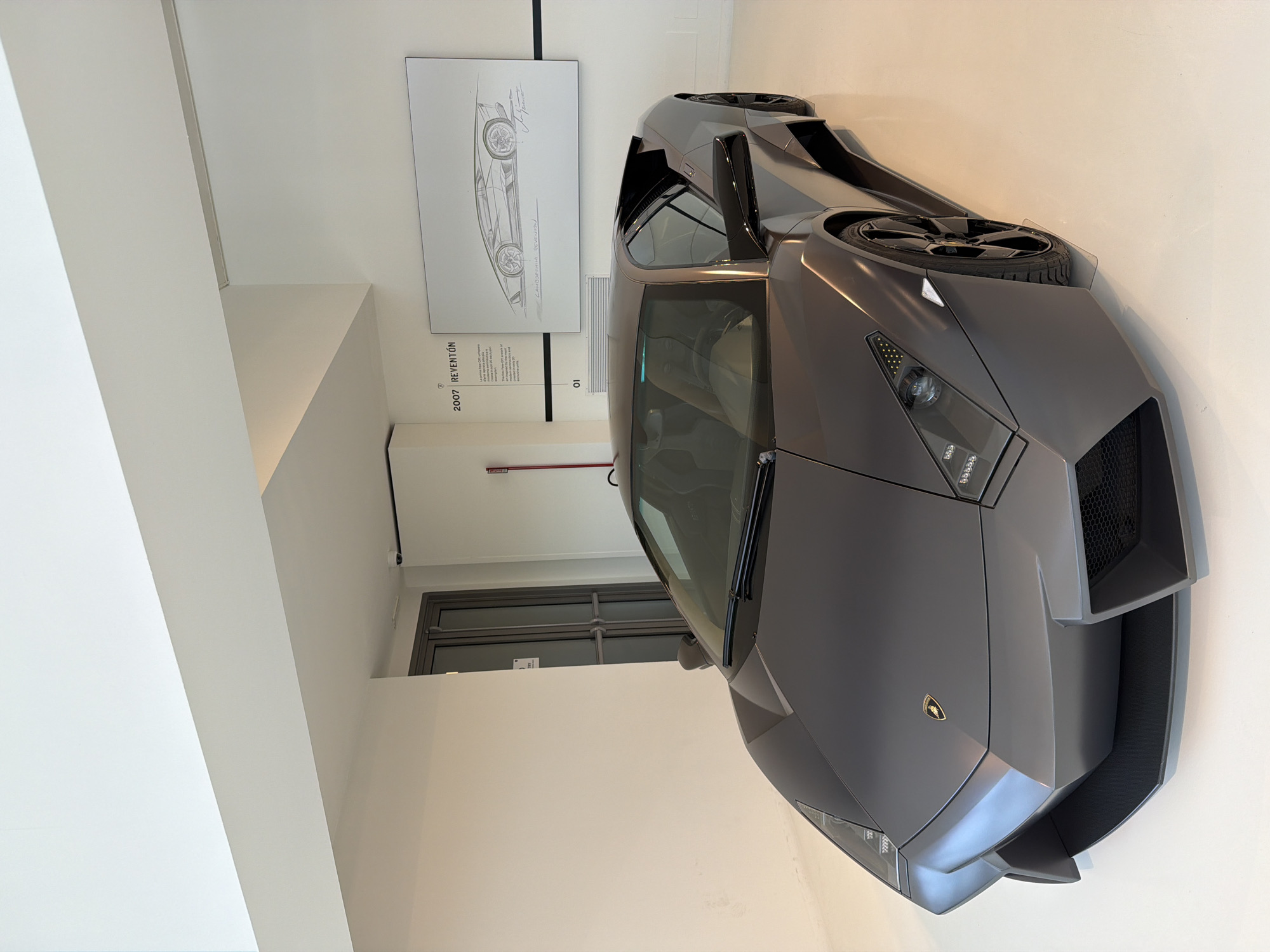 The image displays a dark gray Lamborghini Reventón parked on a polished white floor in what appears to be a showroom or museum. The car is angled slightly towards the viewer, with its front prominently featured. The vehicle exhibits sharp, angular lines characteristic of its design, with visible headlights, a prominent front grille, and a low-profile stance. The Lamborghini logo is visible on the hood.
To the left of the car, a closed glass-paned door is visible. To the right of the car and mounted on the wall is a framed sketch of the Lamborghini Reventón, accompanied by text that reads "2007 REVENTÓN". A small plaque with the number "01" is also visible below the sketch. The walls are a clean, white, and the ceiling is illuminated with bright, recessed lighting, creating a sterile and modern environment. The overall scene suggests a display of the automotive exhibit, possibly in an indoor setting within the city of Maggi, Italy. There are no people present in the image.